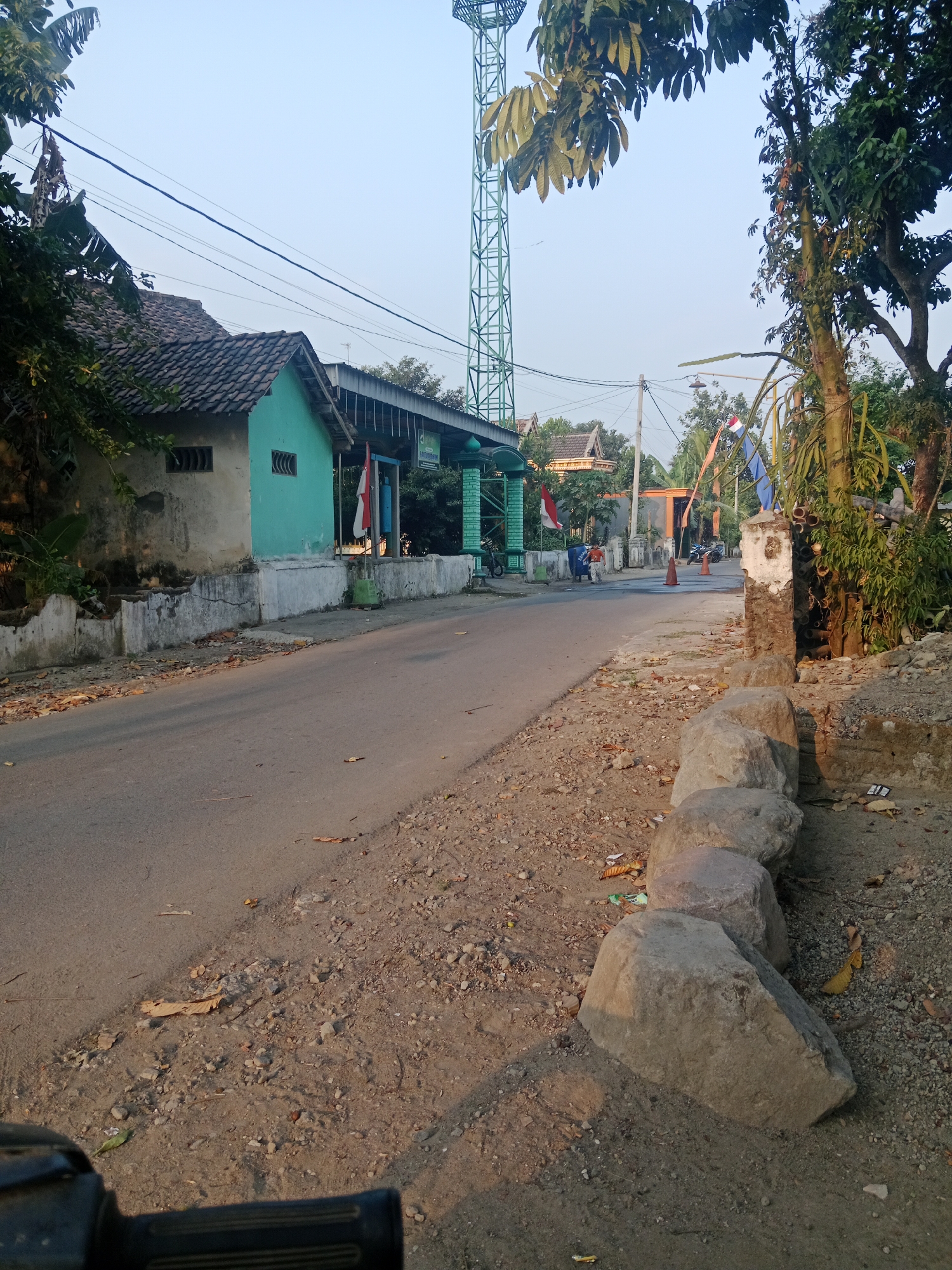 The image depicts a street scene in Ringinrejo, Indonesia. In the foreground, a paved road runs diagonally from the bottom left to the middle right of the frame. The roadside on the right is unpaved and covered with gravel, small stones, and some fallen leaves. Several large, grey rocks are arranged in a line along this edge of the road. On the left side of the road, a row of buildings lines the street. The closest building is a small, weathered structure painted teal, with a dark tiled roof. Further down the street, there are more buildings, some with green accents and white trim. Indonesian flags are visible, attached to poles outside some of the buildings. A tall, green, lattice-like tower, possibly a communication tower, stands prominently in the background. Trees and foliage are present throughout the scene, with a large tree to the right of the frame, its branches extending over the road. The sky is clear and a pale blue. The overall impression is that of a quiet, rural or suburban street.