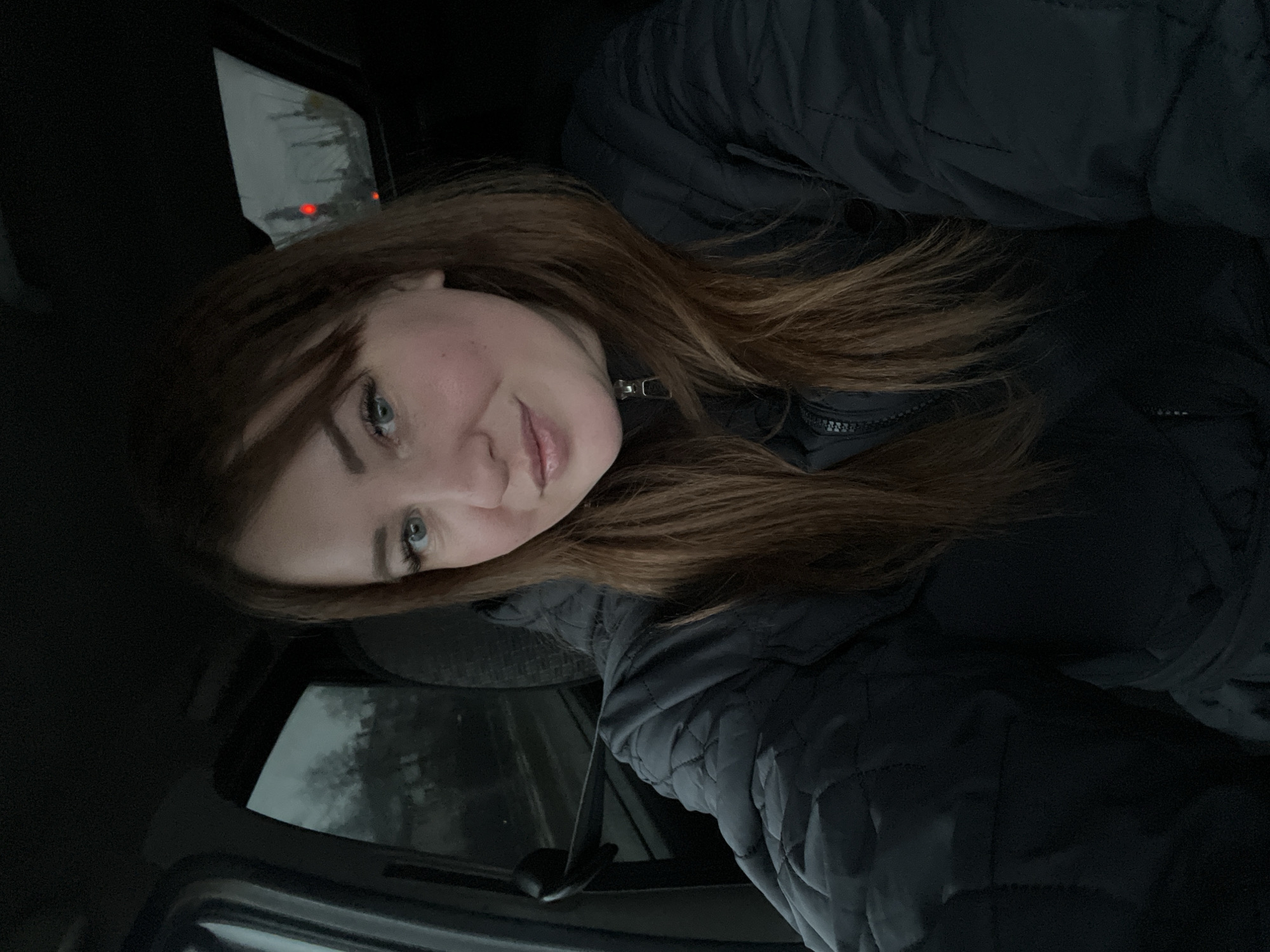 A woman with long, auburn hair is in a vehicle. She is wearing a black jacket. Outside the vehicle's window, it appears to be a cloudy or overcast day. A red light is visible outside the window. Based on metadata, the location is Chaikovsky, Russia.