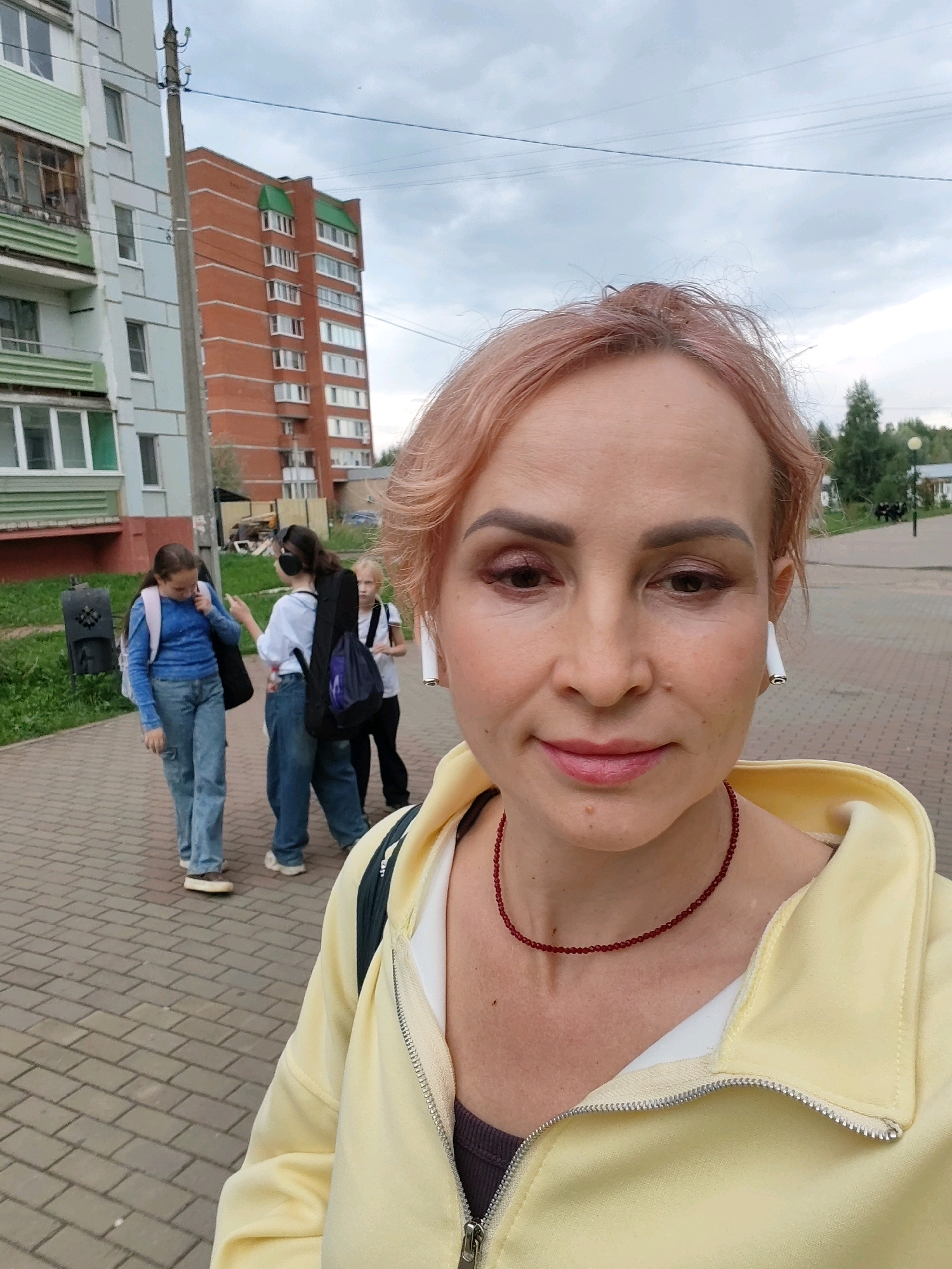 A selfie of a woman with pink hair, wearing a yellow hooded jacket and a beaded necklace, is taken outdoors in Chaikovsky, Russia. In the background, several girls with backpacks walk on a brick-paved sidewalk. Behind them, there are apartment buildings, a street light, and trees. The sky is overcast.