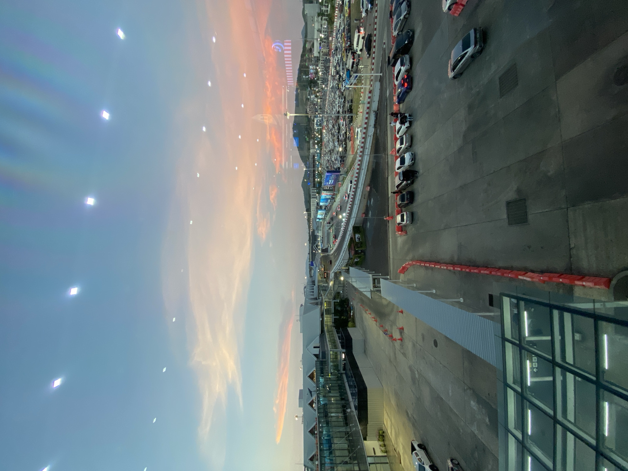 This is an outdoor scene in Mai Khao, Thailand, featuring a mix of structures, vehicles, and the sky at dusk.
In the background, there are mountains and a large parking area packed with cars. There's also a road with traffic. In the middle ground, there is an elevated walkway and surrounding buildings.
The foreground consists of part of a modern building with glass windows reflecting the interior lights. On the ground, near the building, are some parked cars and traffic cones.
The sky is a mix of blues and oranges, possibly indicating sunset or sunrise. There are also what appears to be lights reflected in the window.