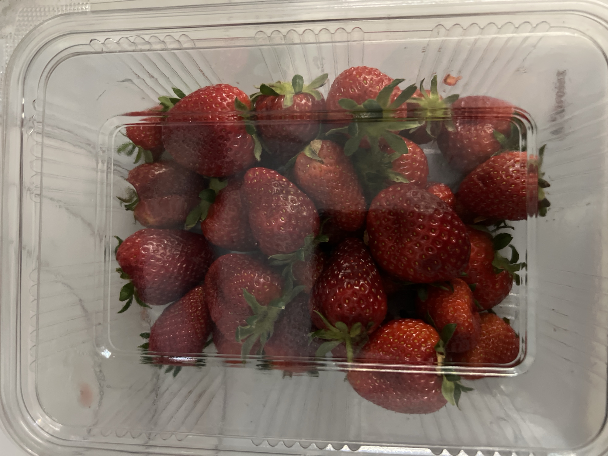 A container of strawberries is on a white surface. The container is clear plastic and rectangular with rounded corners. The strawberries are red and vary in size. The environment is indoors with no people visible. No location details (Burgas, Bulgaria) can be determined from the image alone.