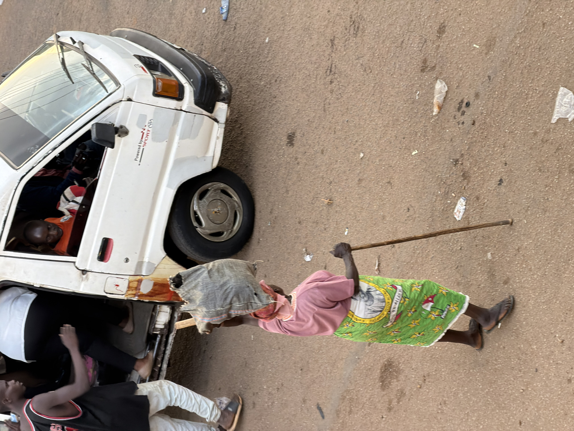 In Bukuru, Nigeria, a woman carries a bag on her head and walks with a stick. She wears a pink shirt and a green skirt with a portrait of a man on it. She has dark skin and is wearing flip-flops. Behind her, people are entering a white van. One person, wearing a black tank top and white pants, stands by the van. Another person, wearing an orange shirt, is visible through the van's window. The ground is a mix of dirt and asphalt with scattered debris. The van has a black bumper and a white body, with some signs of wear and tear.