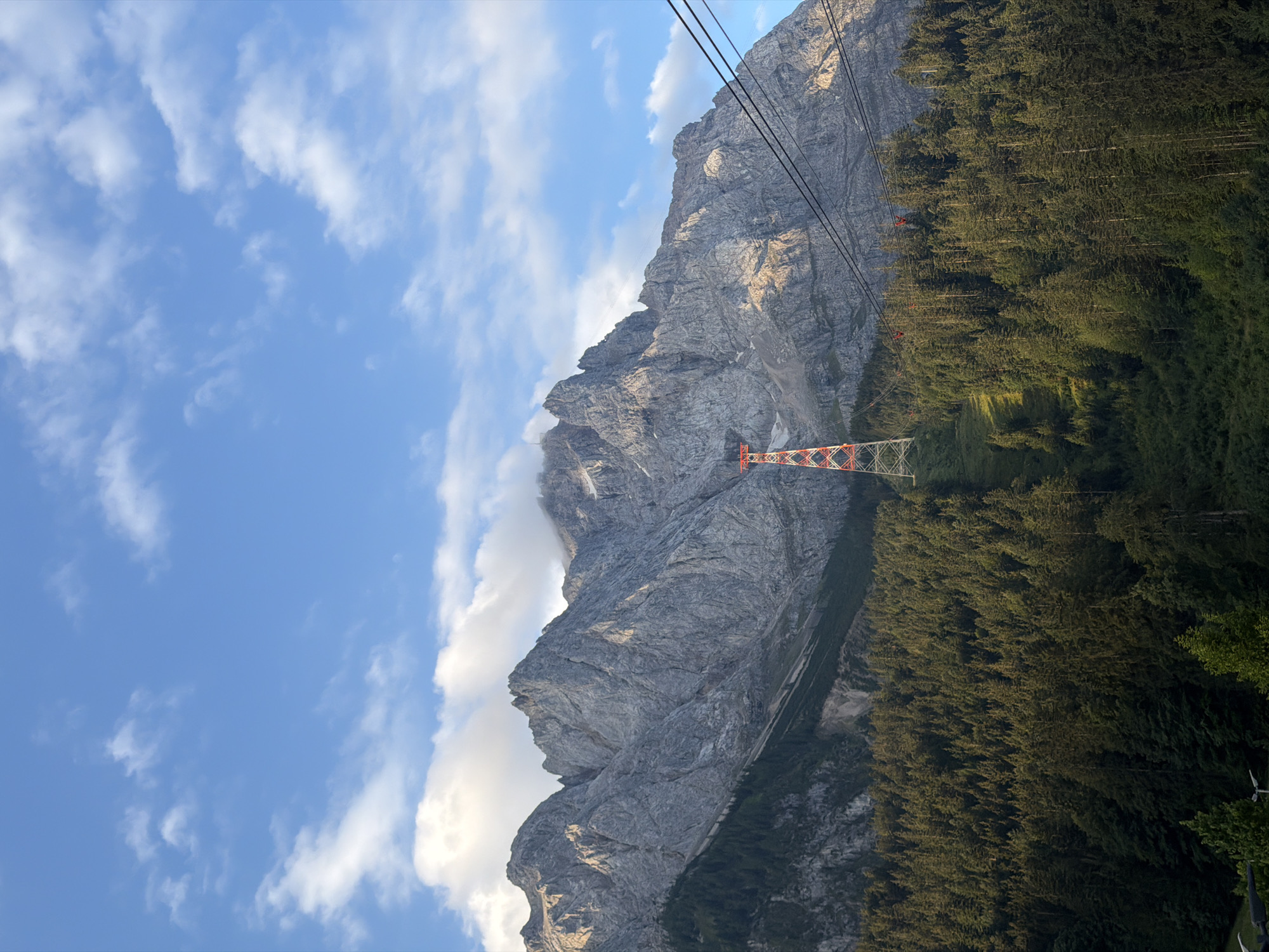 The image shows a panoramic view of the Zugspitze mountain range in Grainau, Germany, under a partially cloudy blue sky. In the foreground, a dense forest of evergreen trees covers a steep slope leading up to the mountains. A tall, red and white pylon, part of a cable car system, stands prominently in the center of the image, ascending the slope through the trees. Several black cables stretch diagonally across the frame from right to left, suggesting the path of the cable car. The rugged, gray rocky faces of the Zugspitze mountains dominate the background, with some snow patches visible on the higher elevations. The sunlight casts shadows and highlights on the mountain slopes, indicating it is either early morning or late afternoon. There are no people visible in the image.
