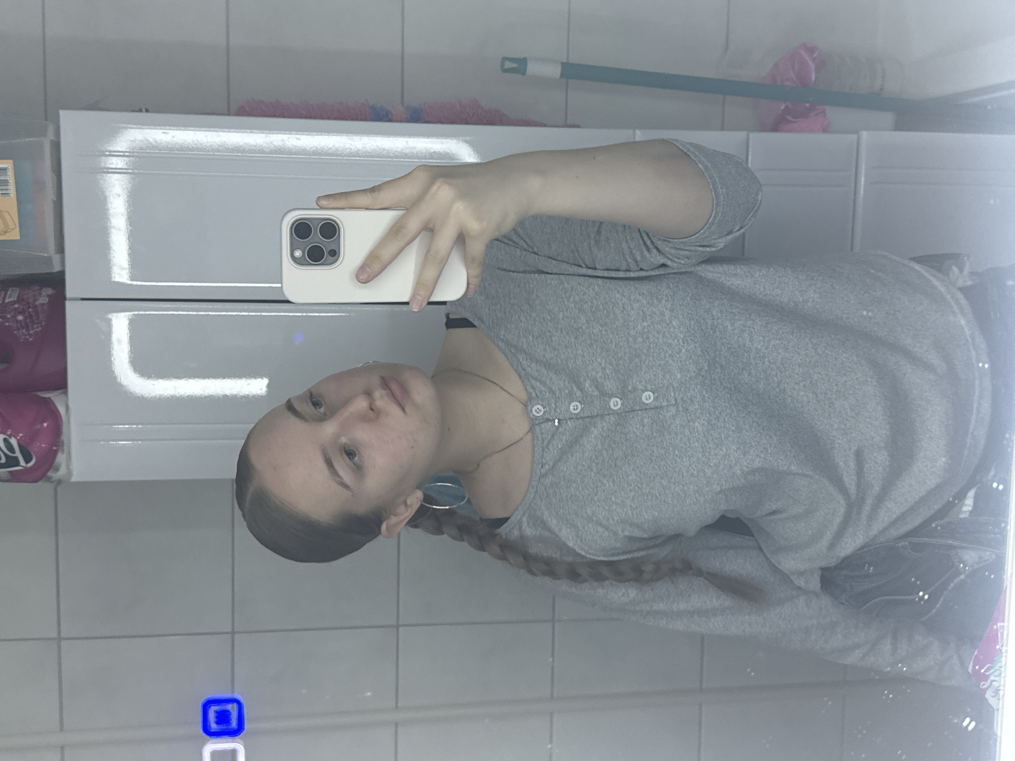 A young female is depicted in a mirror selfie, looking slightly to her right. Her light brown hair is pulled back tightly from her face and styled into a single braid draped over her left shoulder. She wears a grey long-sleeved Henley-style shirt with a button placket and a thin silver chain necklace with a small pendant. A large silver hoop earring is visible in her left ear. Her right hand holds a light-colored smartphone, revealing its triple-lens camera module, positioned to capture her reflection. The background consists of light grey square tiles. Above her reflected head, a light-colored two-door cabinet is mounted, with a pink object and a clear plastic storage container on its top surface. To the upper left of her reflection, a digital display shows illuminated segments and a blue square icon. To the right side of the image, a light blue mop with a white handle and a pink fluffy object are propped against the wall.