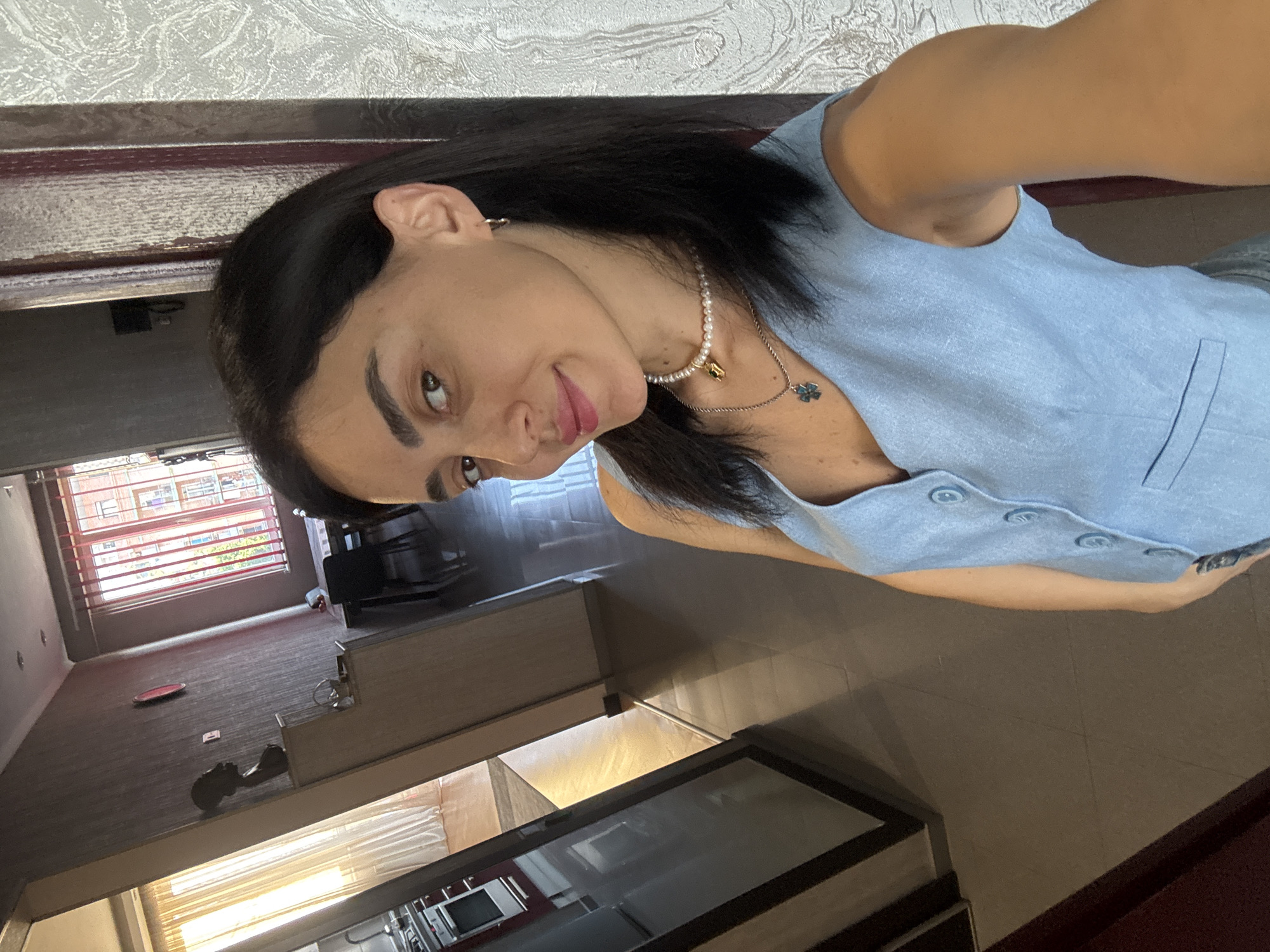 This is a selfie taken by a person with dark hair and brown eyes, wearing a light blue sleeveless top and a pearl necklace. The person is smiling slightly. The background appears to be an indoor setting with patterned wallpaper on the upper wall and a wooden border. Below the wallpaper, the scene shows a room with a dark-colored floor. In the distance, there's a window with horizontal blinds, revealing buildings outside. There are also some furniture items visible, including what looks like a desk or counter area and some shelves. The lighting suggests it could be daytime. The image is oriented sideways.