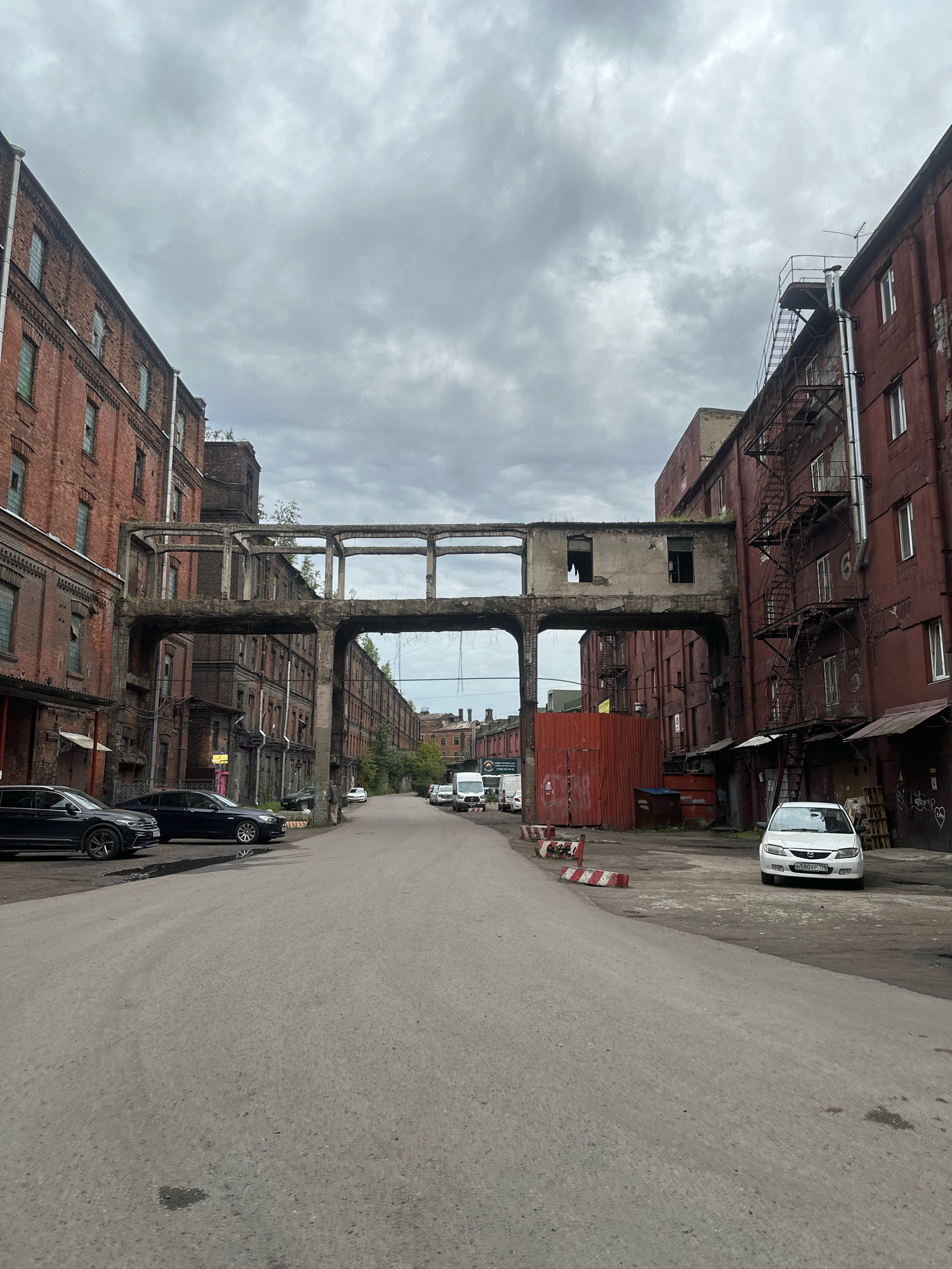 The image depicts an industrial area with aged, brick buildings, likely a former factory complex, located in Saint Petersburg, Russia. The buildings are several stories high, with numerous windows, many of which appear broken or boarded up. A concrete skybridge, showing signs of disrepair, connects two of the buildings across a paved roadway. Red metal barriers are placed along the side of the road, indicating some form of construction or traffic management. Several cars are parked or driving on the road, including a white hatchback in the foreground on the right and two dark sedans on the left. The sky is overcast with gray clouds. The overall impression is one of a historic, possibly neglected, industrial district in the city.