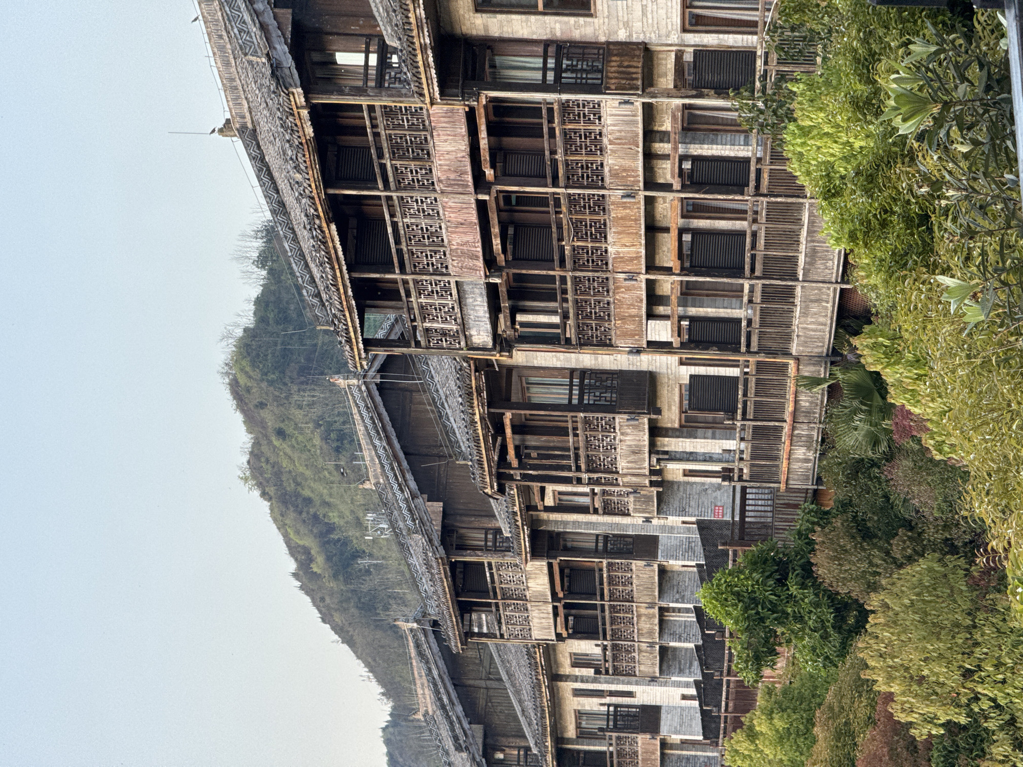 The image shows a multi-story wooden building complex in Zhuoshui, China, situated on a hillside. The building features intricate latticework on its facade and layered, tiled roofs. The complex is surrounded by lush green vegetation, and a mountain rises in the background under a pale, overcast sky.