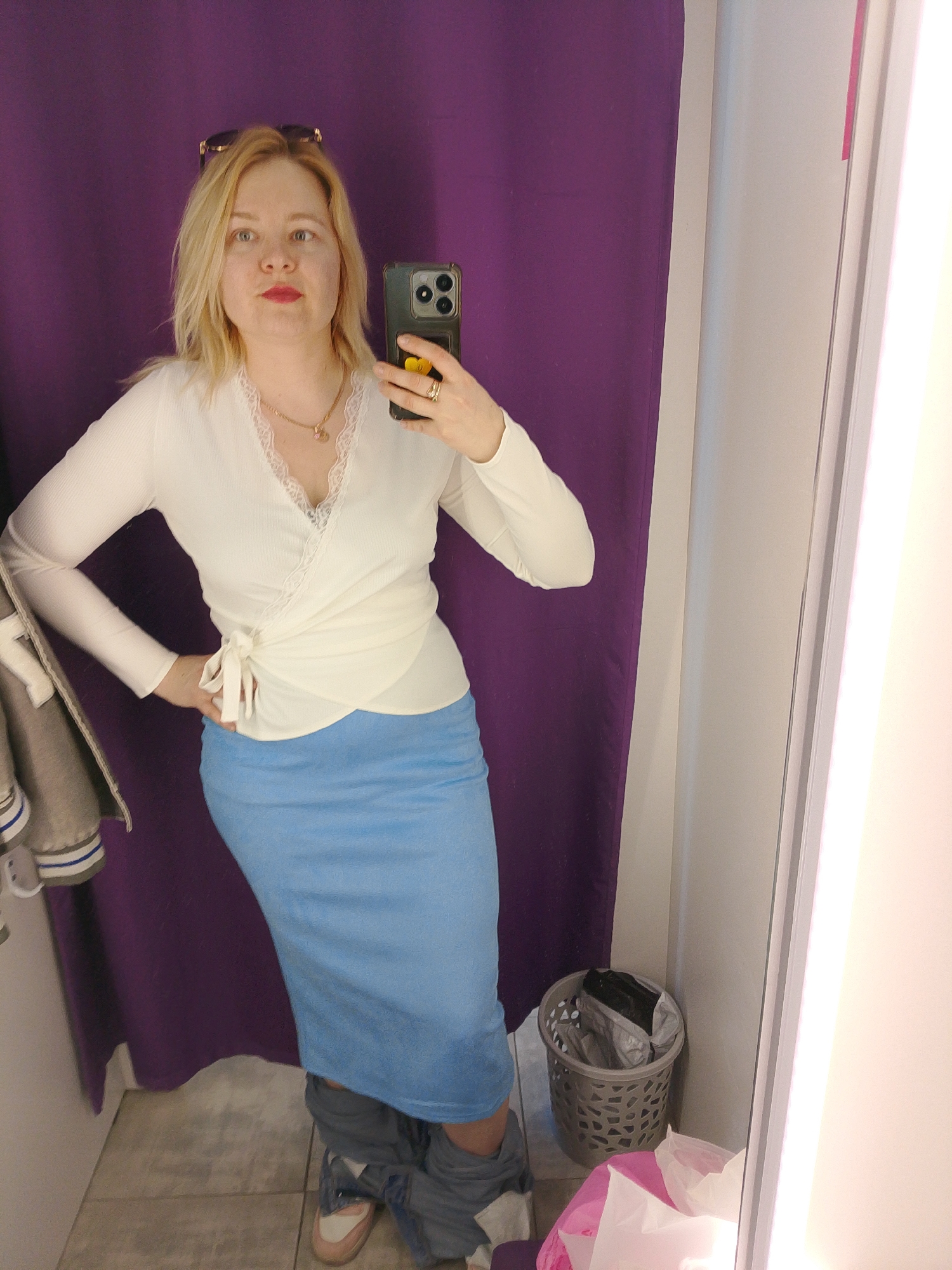 A woman is taking a selfie in a dressing room. The woman has blonde hair and is wearing sunglasses on her head, a white long-sleeved wrap top with lace trim, and a light blue skirt. Her jeans are around her ankles. She is holding a smartphone in her right hand to take the photo. The background consists of a purple curtain and a dressing room mirror. A grey wastebasket containing discarded clothing tags and paper is visible near the bottom right corner of the image. A gray jacket is hanging inside of the dressing room. The location is potentially in Izhevsk, Russia.
