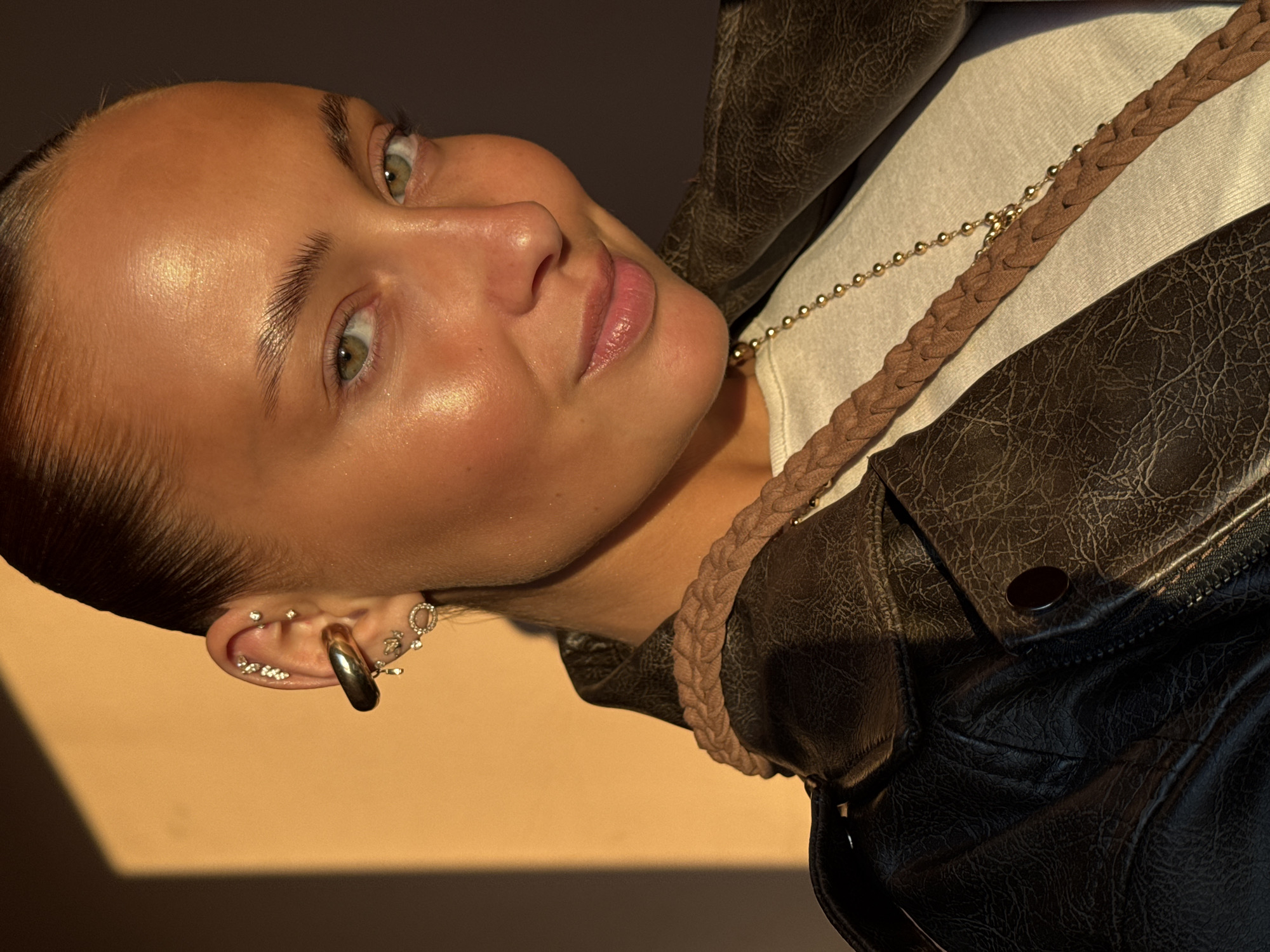 This is a close-up, angled portrait of a young person, likely female, captured during the golden hour, suggesting either sunrise or sunset. The subject has a radiant, tanned complexion, accentuated by the warm, directional sunlight that creates highlights on her forehead, cheekbones, and lips. Her eyes are a striking light green or hazel, and she has neatly groomed eyebrows. Her hair appears to be pulled back tightly, emphasizing her facial features. A soft, pleasant smile is visible on her lips, conveying a sense of contentment or ease.
She is wearing a dark, possibly brown or black, faux leather jacket with a distressed, textured finish. Underneath, a light-colored (off-white or cream) top is visible. Her left ear is adorned with multiple piercings: a prominent, thick, gold-toned hoop earring in the lobe, and several smaller, sparkling stud earrings (likely diamonds or cubic zirconia) along the helix and upper ear, arranged in clusters or individual points. Around her neck, she wears a delicate gold-toned chain necklace with small spherical beads, alongside a wider, braided light brown or tan strap that also features a subtle golden chain woven into it, possibly an element of a bag or another accessory.
The background is minimalist, consisting of a warm, light-toned surface that transitions into a darker, shadowed area towards the lower left of the frame. The strong, low-angle sunlight casts a distinct, sharp shadow, further emphasizing the golden hour setting. The activity is posing for a portrait. No specific location cues are visible within the frame that would explicitly indicate Chaikovsky, Russia. There is no visible text.