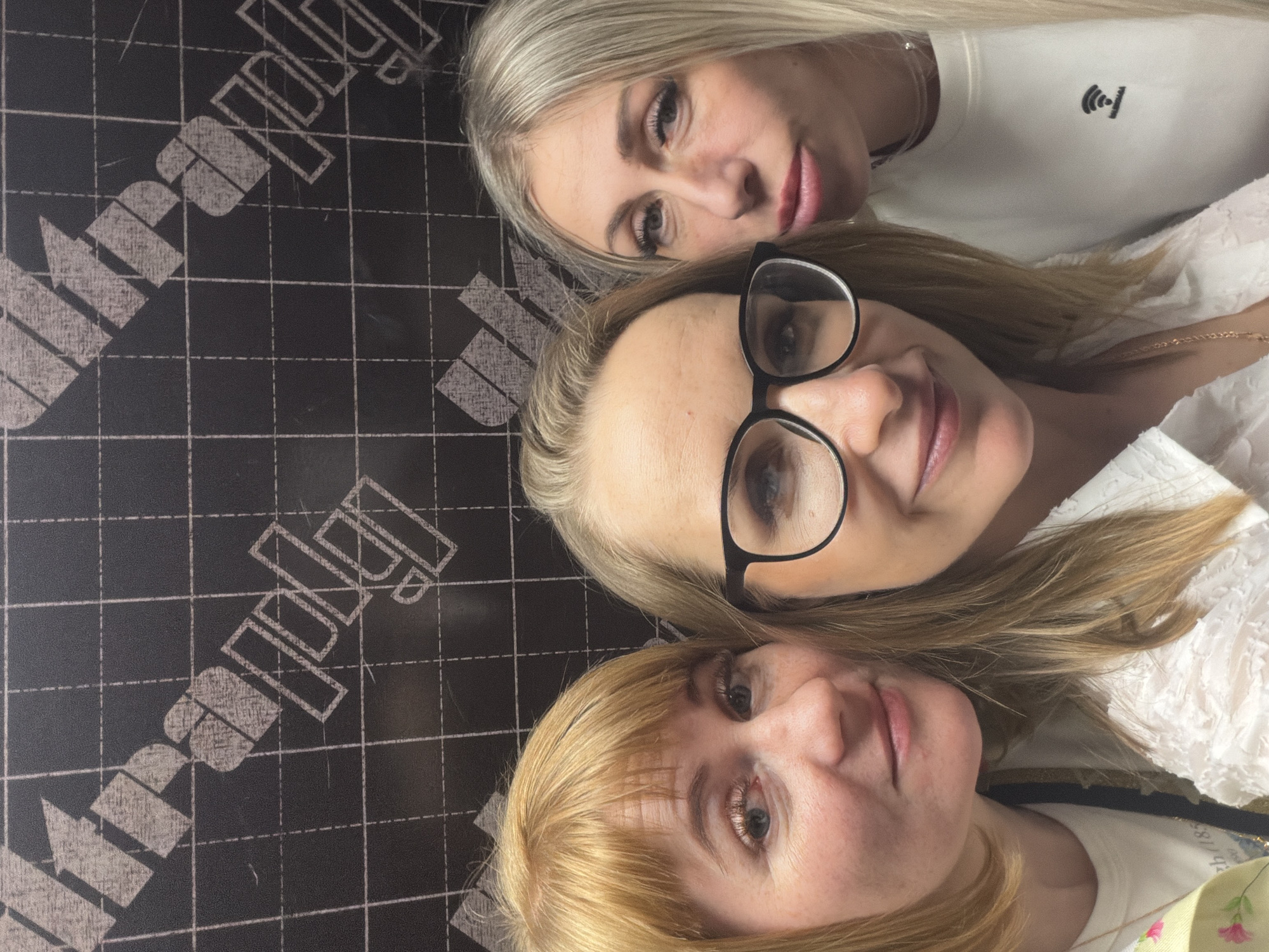 Three fair-skinned women pose closely together, possibly for a selfie. The woman at the top has blonde hair and is wearing a white shirt with a small black logo. The woman in the middle has blonde hair and is wearing black-rimmed glasses. The woman at the bottom has blonde hair with bangs. They are indoors, with a black textured surface with a white grid pattern and a white logo on it visible in the background. This event is taking place in the city of Khimki, Russia.