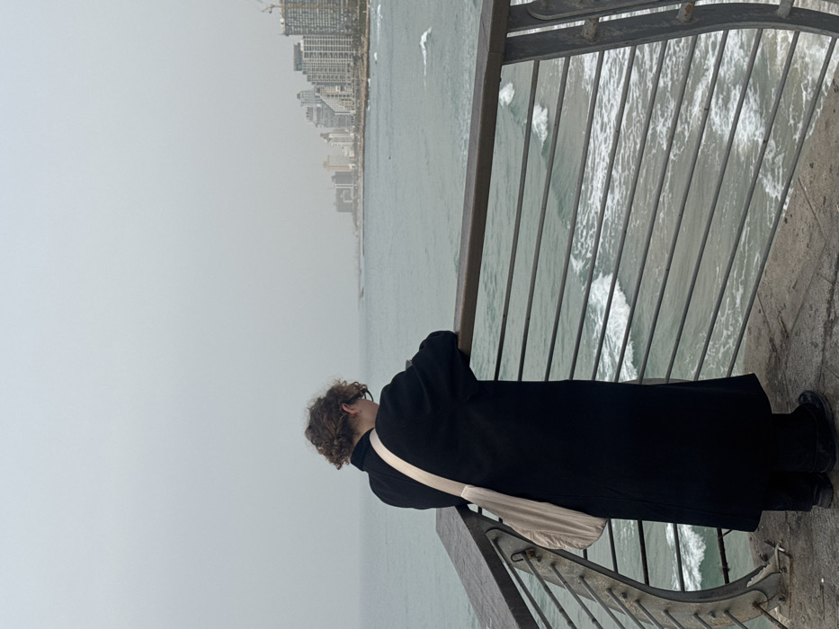 A person is depicted from behind, angled slightly left, standing on a pier or promenade and gazing out at the Mediterranean Sea. The individual has medium-length curly brown hair and wears glasses, a long, dark (likely black) coat, dark trousers, and dark shoes. A light-beige fabric bag with a wide strap is worn across their left shoulder. They are leaning forward with their forearms resting on a railing that features a dark wooden top bar and multiple horizontal metal bars. The sea below is choppy with visible white-capped waves. The sky above is uniformly overcast and grey. In the distance, along the coastline, the distinctive skyline of Tel-Aviv, Israel, is visible, characterized by numerous modern high-rise buildings and several operational construction cranes. The scene suggests a vantage point from the Jaffa Port area looking north towards central Tel-Aviv.