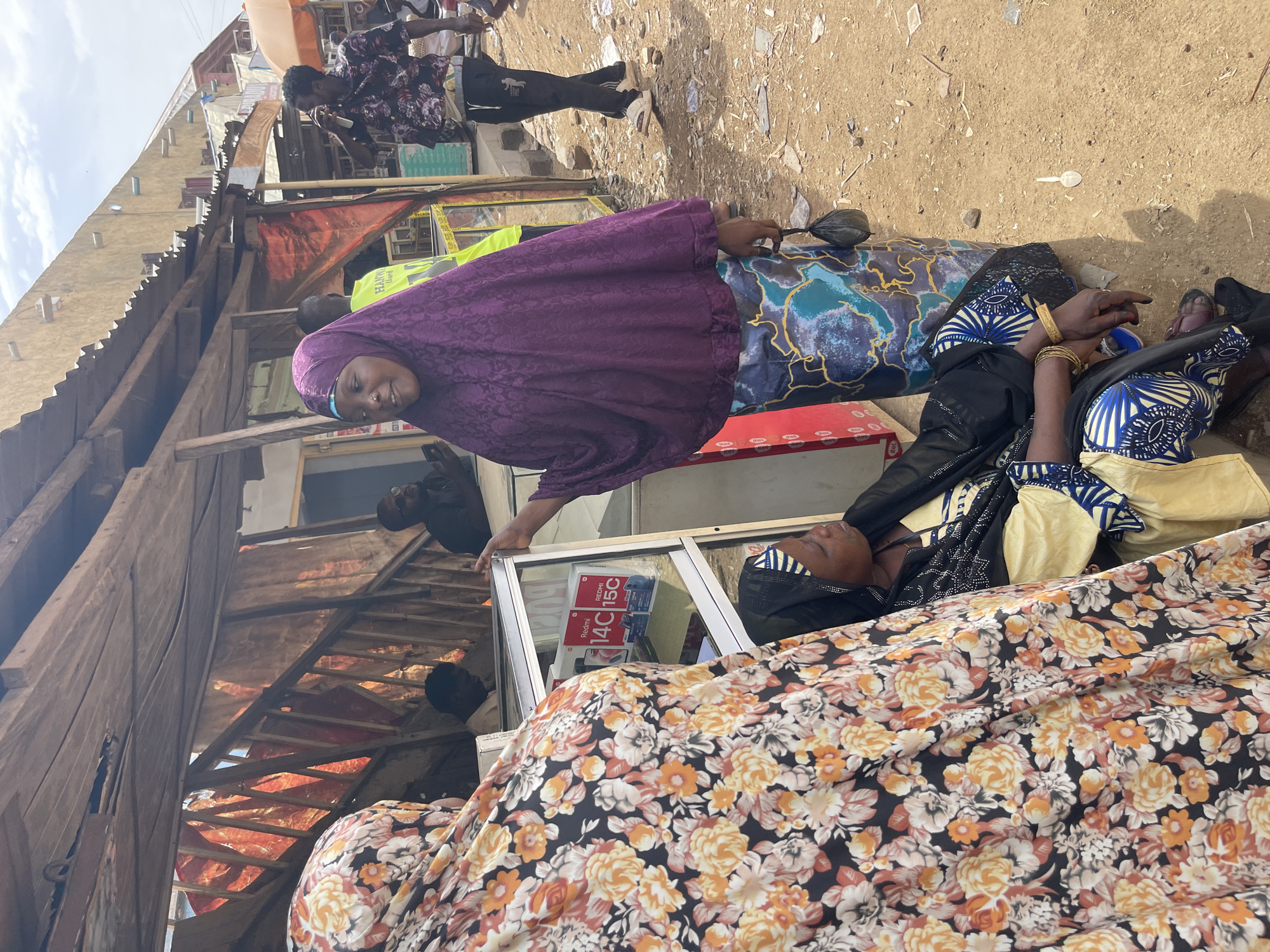 Two women are seated on the ground in what appears to be a market or commercial area in Chikaji, Nigeria. One woman wears a purple head covering and floral patterned clothing; the other wears clothing with blue and black patterns. A glass-enclosed kiosk displays "Redmi 14C 15C" and cell phones. Other people are visible in the background; the environment is dusty and contains corrugated metal structures.