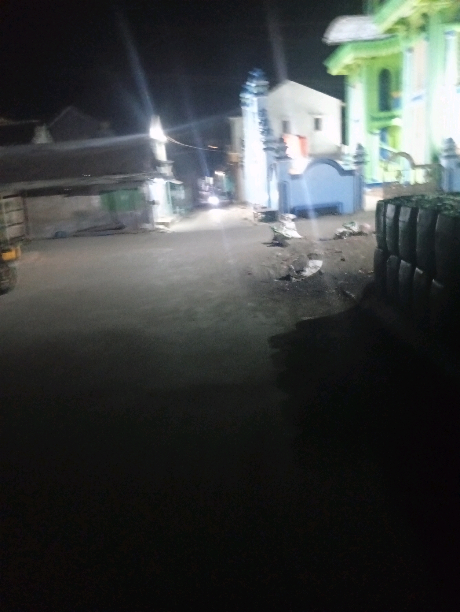The image shows a street scene at night in Banowan, Indonesia. A building with a green facade and a white roof is visible on the right, possibly a mosque or a similar structure. To the left of this building, there is a white building with blue accents. In the foreground, stacks of what appear to be dark, cylindrical objects are piled up. The street is paved and illuminated by streetlights, creating long light streaks due to the low light conditions. A vehicle is partially visible on the far left. The environment appears to be a residential or commercial area in a town or city.