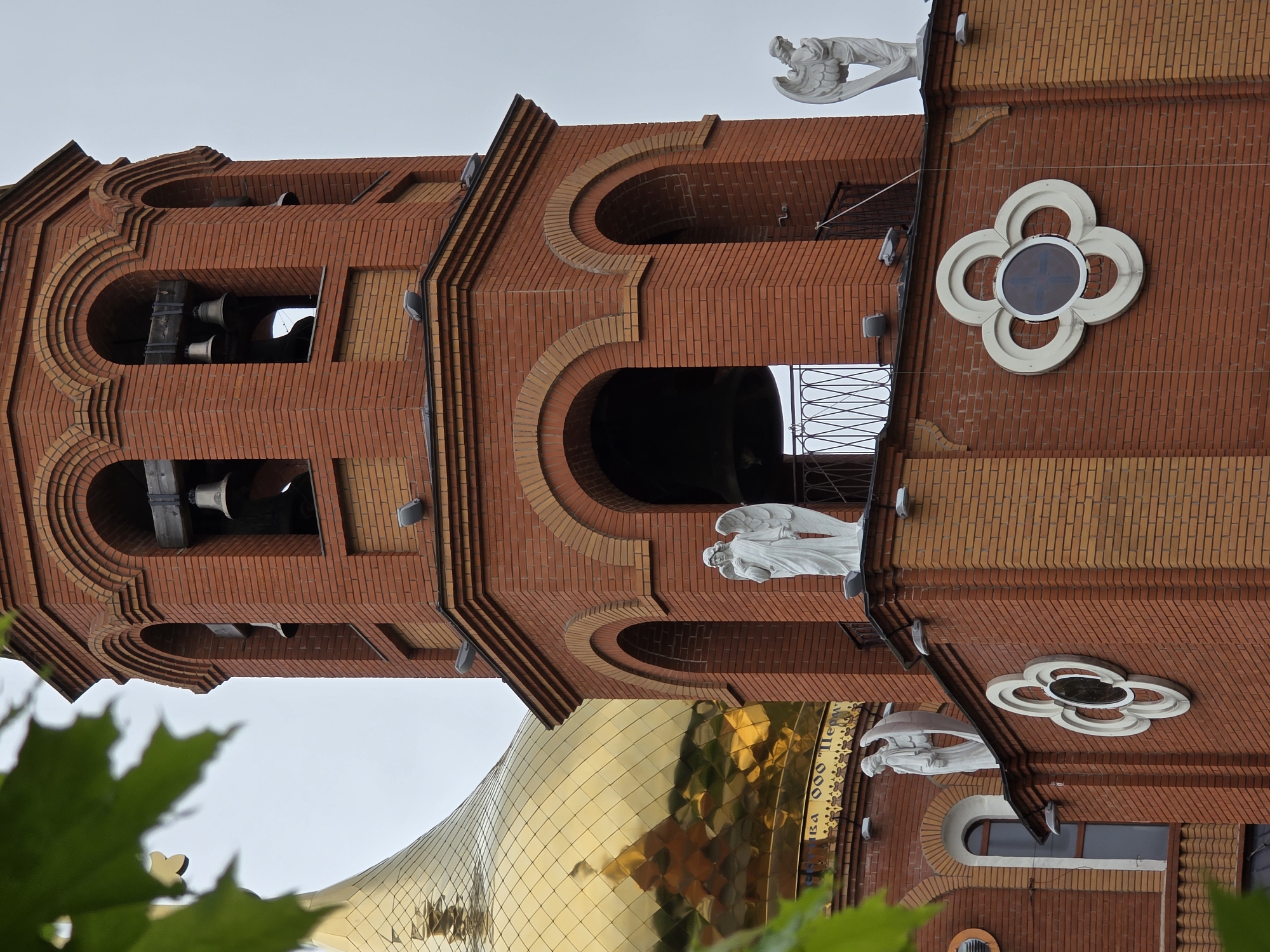 The image shows a brick building with a golden dome, likely a church, in Chaikovsky, Russia. The architectural details include arched windows and decorative brickwork. A bell tower with visible bells is present. Statues of angels are positioned on the building. There are also white, flower-shaped decorations on the wall. The perspective is from below, looking upward, with green foliage partially obscuring the view. The sky is visible in the background.
