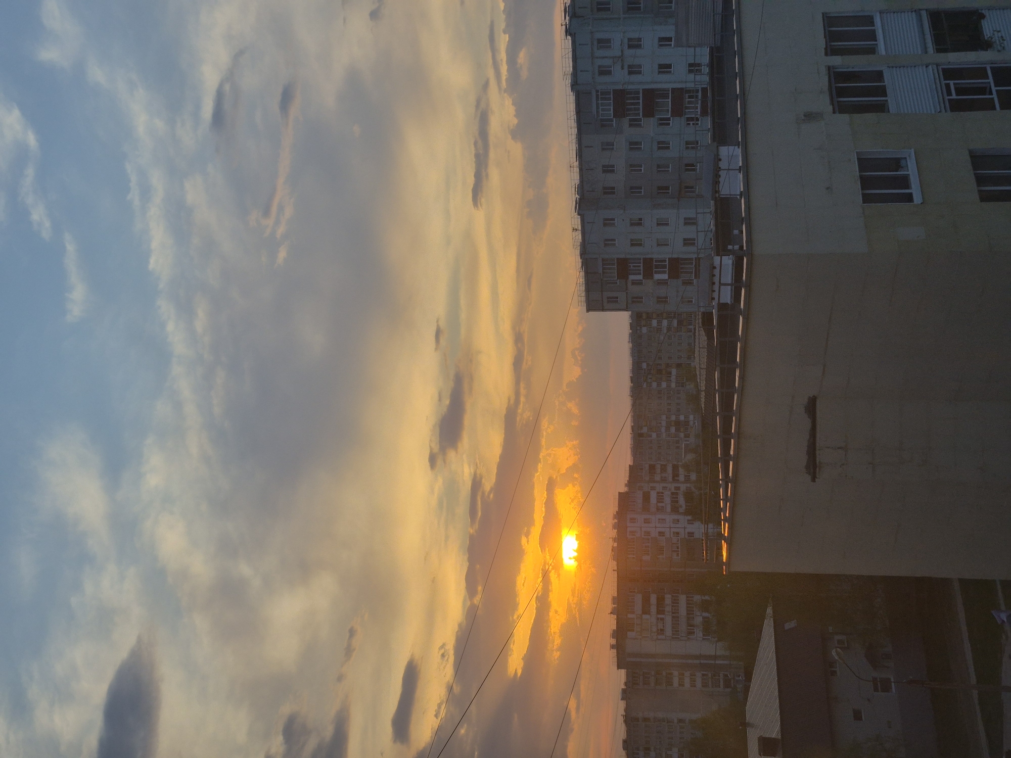 The image depicts a sunset over a cityscape, with the location being Unknown, Unknown. The sky is filled with clouds, ranging in color from blue to orange and yellow due to the setting sun. The sun itself is visible, appearing as a bright yellow orb partially obscured by the clouds. In the foreground, there are buildings, including high-rise apartment blocks, with numerous windows visible. Some of the buildings are under construction or renovation, as evidenced by scaffolding on the exterior. Power lines are also visible, cutting across the sky. The overall scene captures the urban environment with the natural beauty of a sunset.