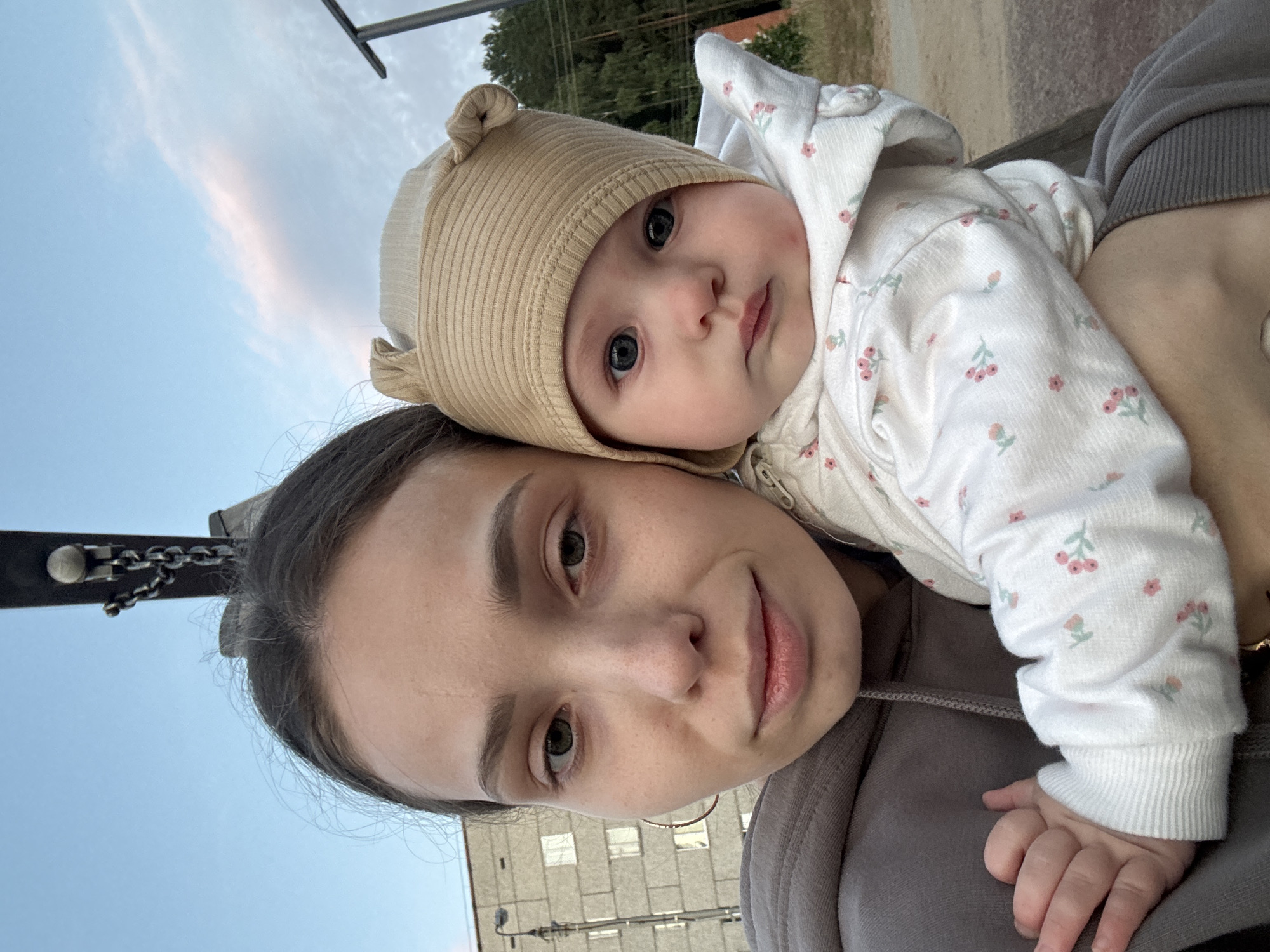 A young woman with light skin, dark hair pulled back, and green eyes is holding a baby. The woman is wearing a grey hooded sweatshirt. The baby, with light skin and blue eyes, wears a tan hat with ear details and a white onesie with a floral pattern. They are outdoors, with a grey building and utility pole visible in the background to the left. Trees and a dark lamppost are in the background to the right. The sky is partly cloudy. The scene appears to be in a park or recreational area. The city of Noviy, Russia is indicated as the location.