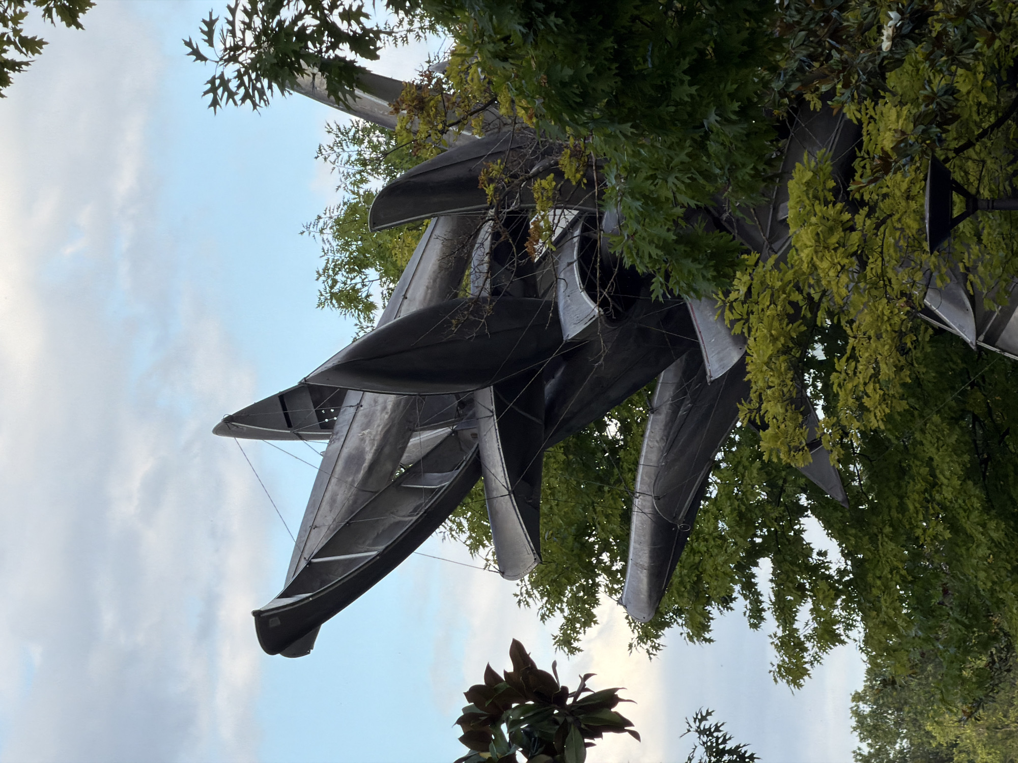 A low-angle, medium shot captures a large, abstract metal sculpture of a geometric, fan-like shape, comprised of overlapping, elongated curved planes. The sculpture is set against a pale blue sky with scattered, wispy clouds. Lush green foliage, including broad leaves and smaller, oak-like leaves, partially obscures the lower portions and sides of the sculpture, with some leaves appearing yellow-green, suggesting early autumn. A dark, cone-shaped light fixture is visible at the bottom center of the frame, integrated amongst the trees. The sculpture appears to be outdoors, in an urban park or public space in Paris, France, given the presence of mature trees and what appears to be a street lamp. No people are visible. The overall lighting is soft, indicating it is daytime.