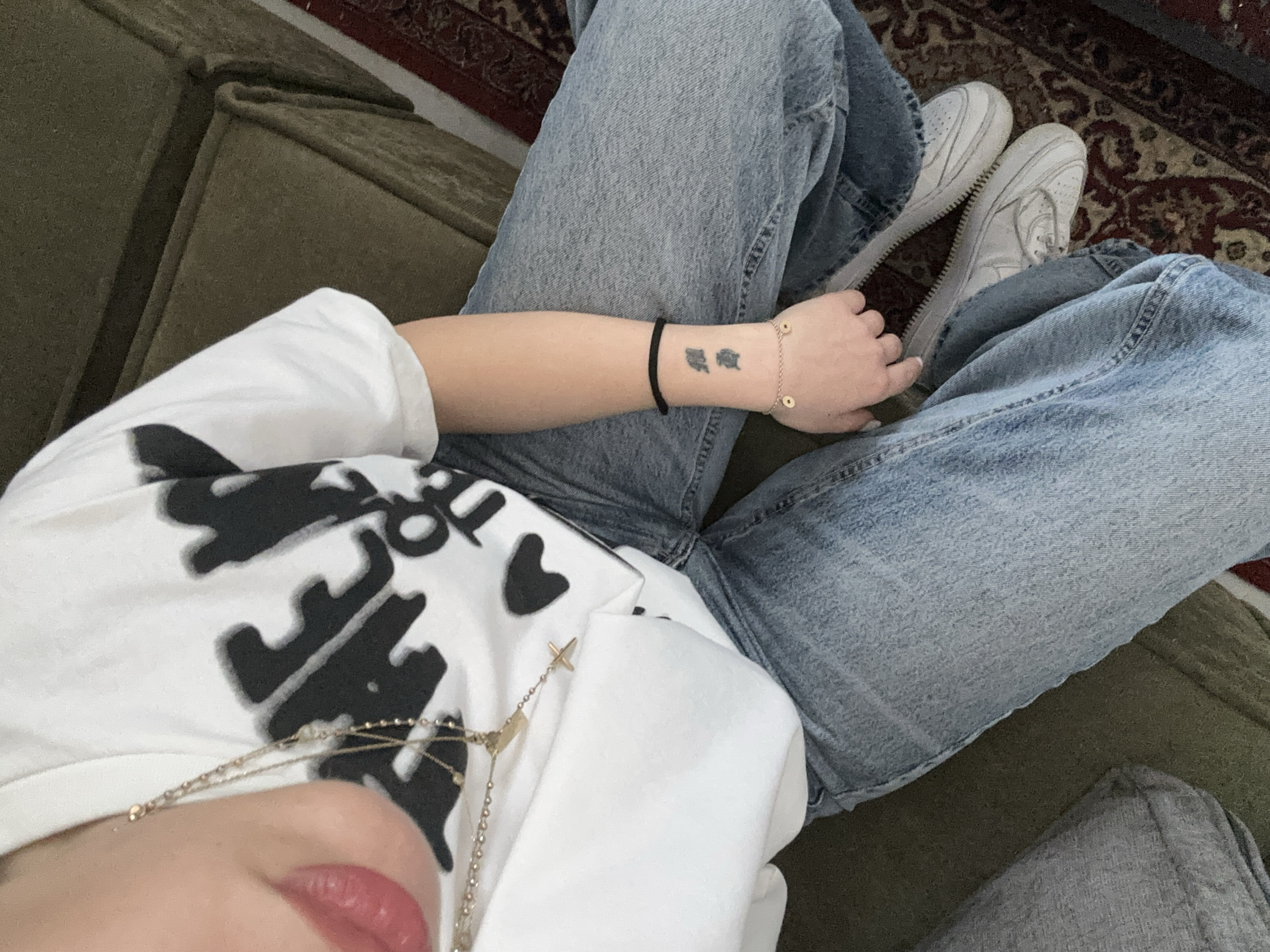 A top-down interior shot depicts an individual seated on a dark olive-green upholstered sofa or armchair. The subject, whose face is not visible, is wearing a white short-sleeved t-shirt with a black, partially obscured graphic design, light-wash blue denim jeans, and white sneakers. A multi-strand gold-colored necklace featuring a cross pendant and a rectangular charm is visible around the subject's neck. The subject's mouth area shows visible red lipstick. On the right wrist, a black elastic band is present, along with a thin gold-colored bracelet adorned with two small circular charms. A tattoo resembling two stylized Asian characters is visible on the inner right forearm. The feet rest on a patterned floor rug, primarily featuring dark red, brown, and cream tones in a traditional motif. The immediate environment suggests an indoor, private setting. No visual information within the image definitively identifies the location as Yerevan, Armenia.