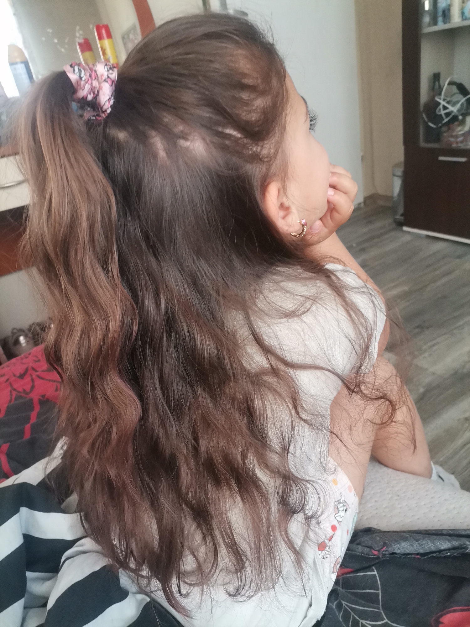 The image displays a young child, likely a girl, viewed from a back and side perspective in an indoor setting. Her long, wavy hair, which is predominantly dark brown with lighter reddish-brown highlights, is partially tied into a high ponytail using a pink patterned scrunchie. A lighter patch or area of less dense hair is noticeable on the scalp behind the scrunchie. The child is wearing a small golden C-shaped hoop earring with a pink stone in her right ear, and her right hand is positioned near her chin or mouth, suggesting contemplation or observation as she looks towards the right side of the frame.
She is dressed in a white top and a patterned diaper featuring small, colorful cartoon-like figures. The child is partially covered by or resting on dark-patterned fabric, possibly bedding or a blanket, with additional black and white striped fabric visible. The background is softly blurred, revealing a domestic interior with a light-colored wall, a dark floor, and a brown cabinet containing indistinct objects. The overall impression is one of quietude. No text is visible within the image, and the time of day is not discernible.