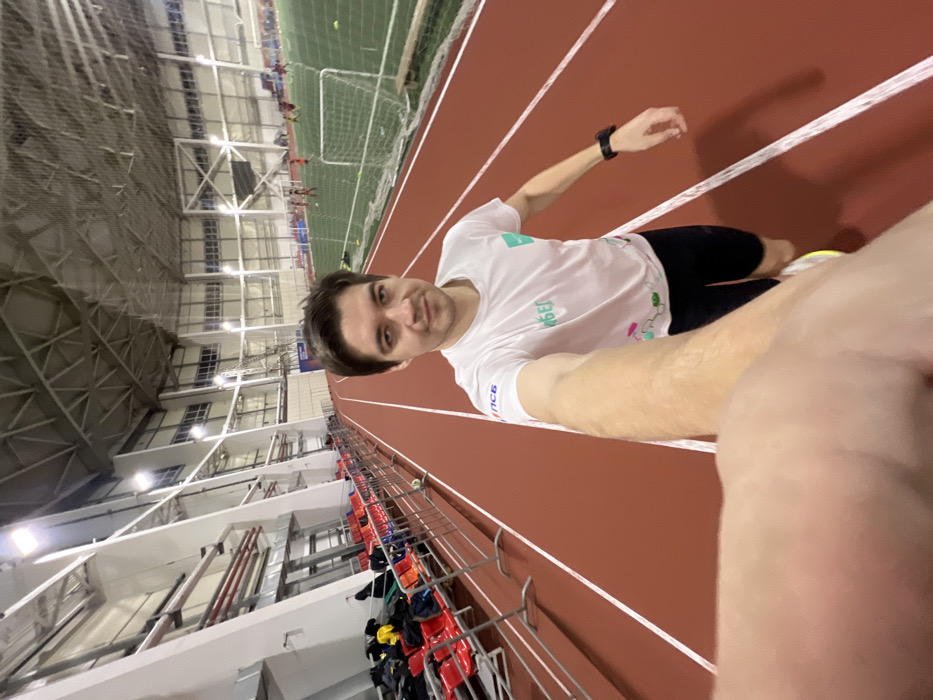 A selfie taken indoors shows a man on a red running track. He wears a white t-shirt and black shorts. A smartwatch is visible on his wrist. The background reveals an indoor stadium with seating and a netted soccer field. The photo appears to be rotated, with the ceiling and stadium structure angled to the left. The man looks toward the camera, holding his arm out for the selfie.