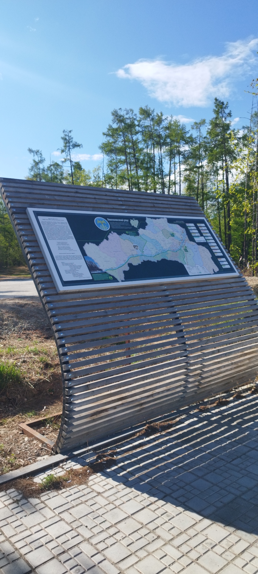 The image shows an outdoor information board in Tekhtiur, Russia. The board, made of horizontal wooden slats arranged in a slightly curved shape, displays a map of the region titled "Хангаласский улус" in Russian. The map is set against a dark background and features a river system, various geographical areas, and smaller informational panels along the right side. The board is positioned on a paved area of light gray interlocking pavers. The background includes a road to the left and a forest with tall, slender trees under a bright blue sky with scattered clouds. The lighting is bright, casting shadows from the wooden slats onto the pavers below. There are no people visible in the scene. The environment appears to be a natural park or recreational area.