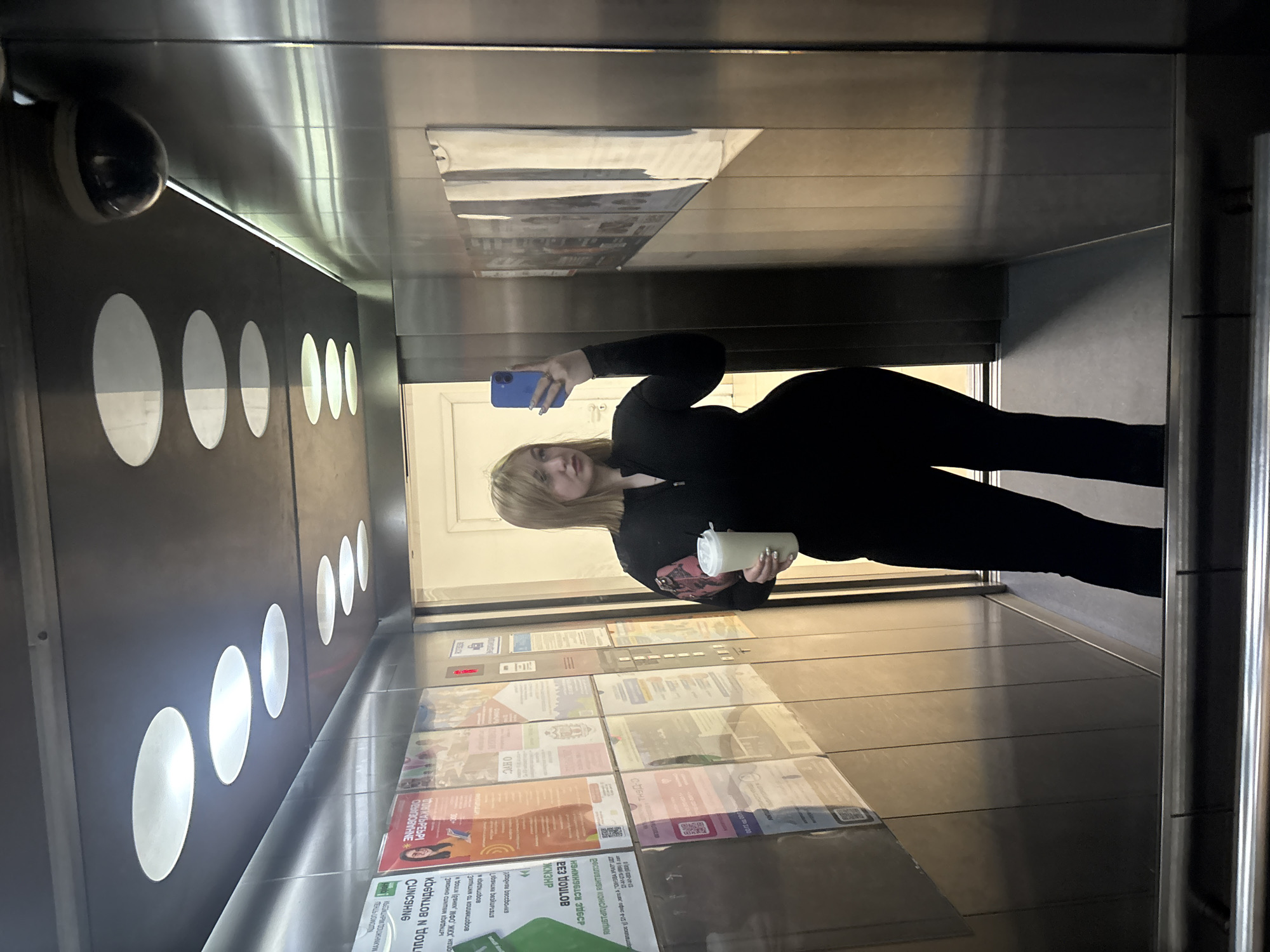 A young woman with blonde hair takes a mirror selfie inside an elevator. She is wearing a black long-sleeved top and black leggings, and is holding a light-colored cup in her right hand. Her left hand is holding a blue smartphone. The elevator interior is modern, with brushed metal walls and a tiled floor. There are several circular, illuminated panels on the left wall. A security camera is visible in the upper left corner. The elevator doors reveal a hallway with posters or advertisements displayed on the wall. The posters contain text in Cyrillic script, including "ОНУС", "PecUUBLHS KOHCAUPLSTING", "РЕЗ НОУЛОВ", "ЖN3HP", "CUNCSHNG", and "NHABAR". The scene appears to be indoors, likely during the daytime.