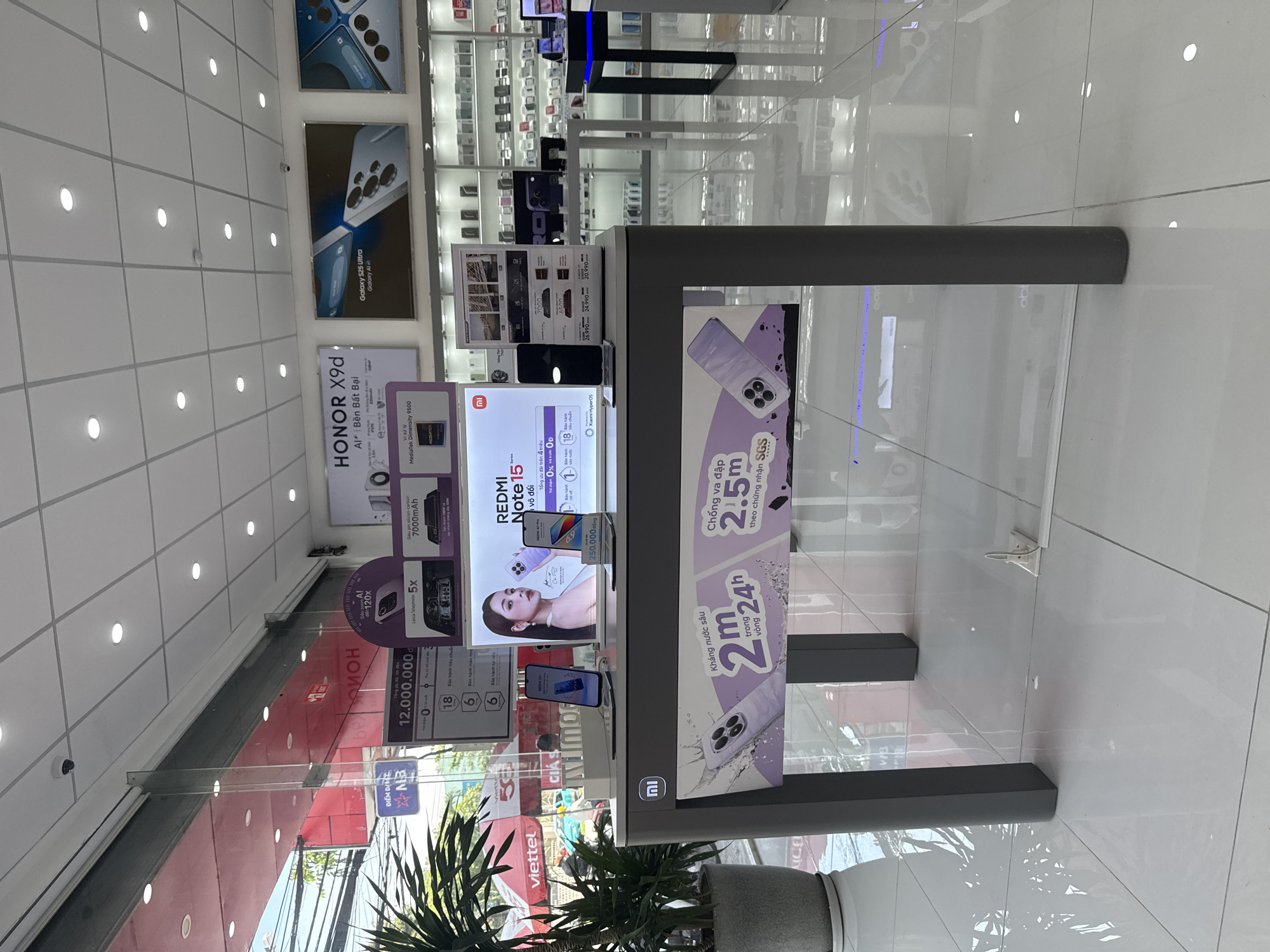 The image depicts the interior of a mobile phone store, likely in Bac Nha Trang Ward, Vietnam, given the Vietnamese text on signage and the presence of a Viettel umbrella outside. The store has a modern, clean aesthetic with white tiled flooring and a white drop ceiling with recessed lighting.
In the foreground, a grey display table with the Xiaomi logo is present, featuring a large banner showcasing a smartphone with a purple gradient design and text in Vietnamese about water resistance and dust protection. Two smartphones are displayed on stands on top of this table.
Behind the display table, a wall of shelves is visible, stocked with various mobile phones. Further back, larger promotional posters are displayed on the walls. One poster advertises "HONOR X9d" and another displays "Galaxy S25 Ultra Galaxy AI". A glass-fronted cabinet or display case is also visible, containing rows of mobile phones. Outside the store, through a large glass window, a street scene with buildings and foliage is partially visible.