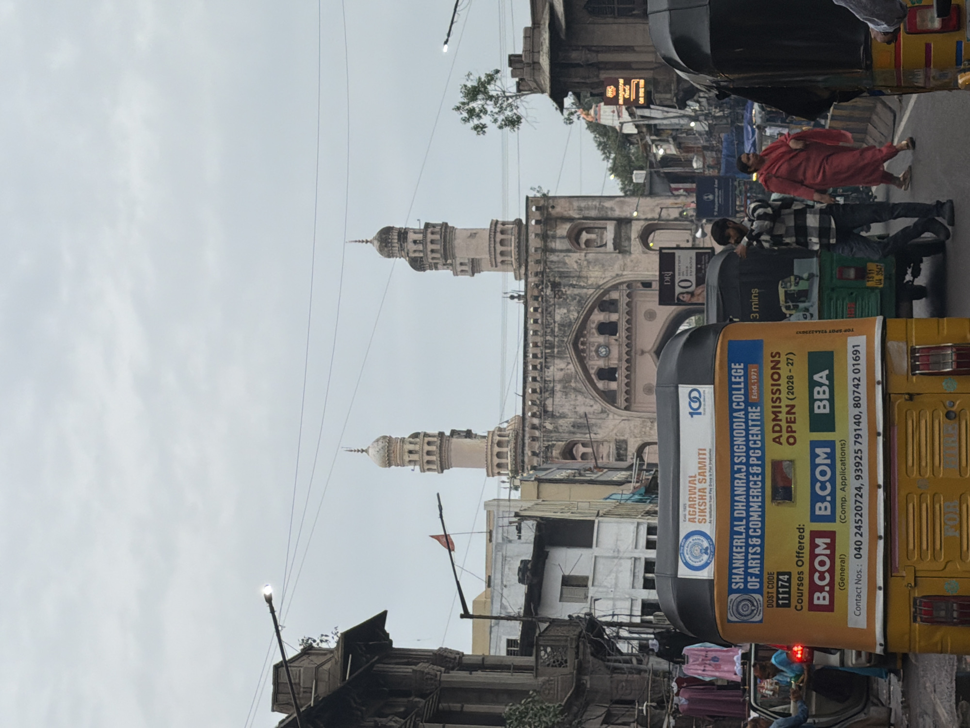 In this full-frame, eye-level shot, a street scene unfolds in Hyderabad, India, with the iconic Charminar monument visible in the background under an overcast sky. A busy street is filled with traffic, including several auto-rickshaws and other vehicles. On the left, an auto-rickshaw displays advertisements for "SHANKERLAL DHANRAJ SIGNODIA COLLEGE OF ARTS & COMMERCE & PG CENTRE," listing courses and contact numbers. Two women and a man are visible near the auto-rickshaws. One woman, wearing a blue top and skirt, stands by a car. Another woman in a red sari walks towards the right of the frame, and a man in a checkered shirt and jeans walks towards the left, gesturing with his right hand. Buildings line the street, some with traditional architecture and balconies. A red flag is visible on a pole in front of one of the buildings. The scene is bustling with activity, characteristic of a busy urban environment.