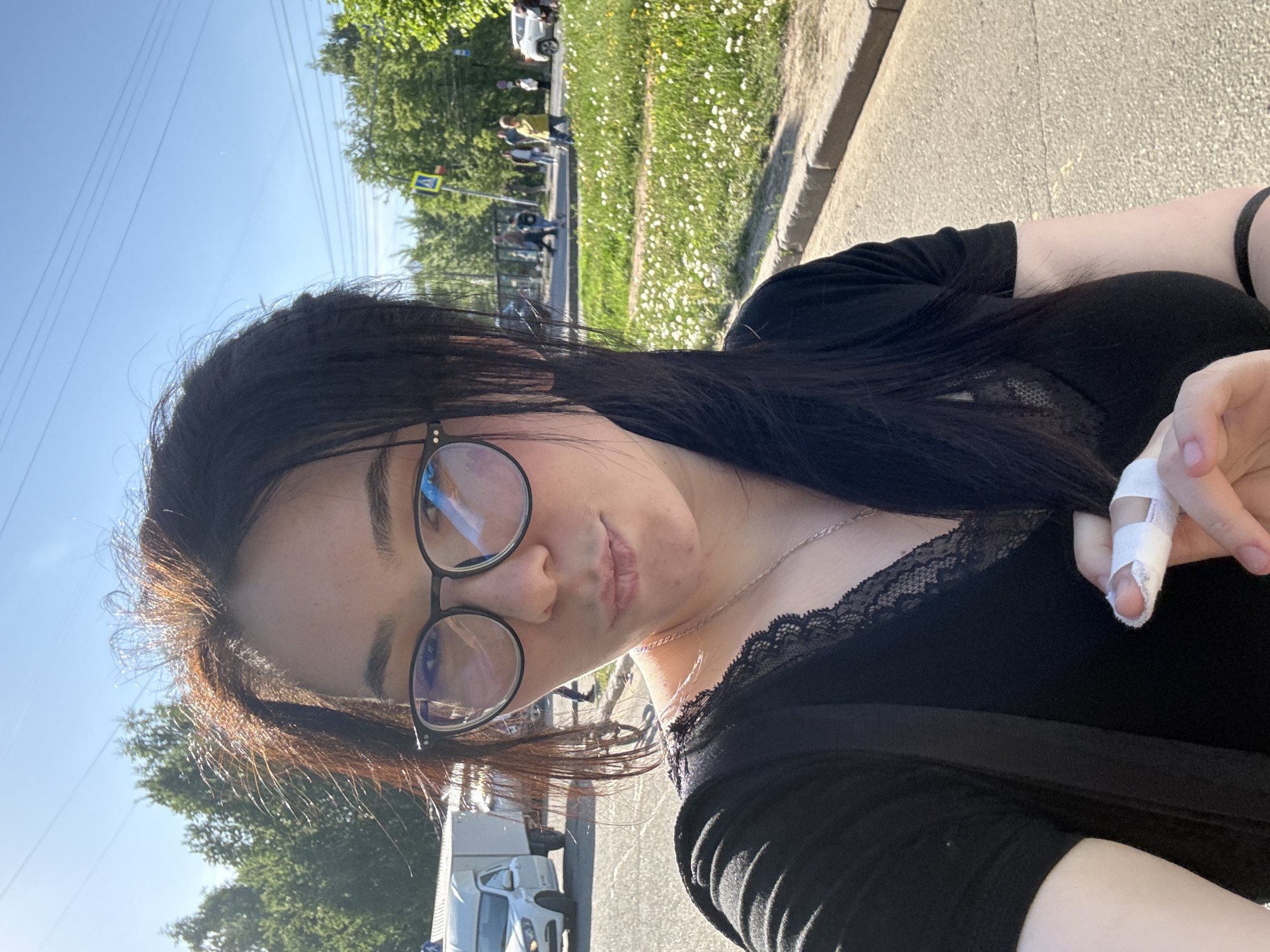 A selfie of a person with dark hair and glasses is oriented vertically. The person wears a black top with lace trim and a silver necklace. A bandage is on the index finger of their right hand. The background includes a sidewalk, a grassy area with white flowers, trees, and a road with cars and pedestrians. Based on visual cues such as the license plates, the location is likely Izhevsk, Russia.