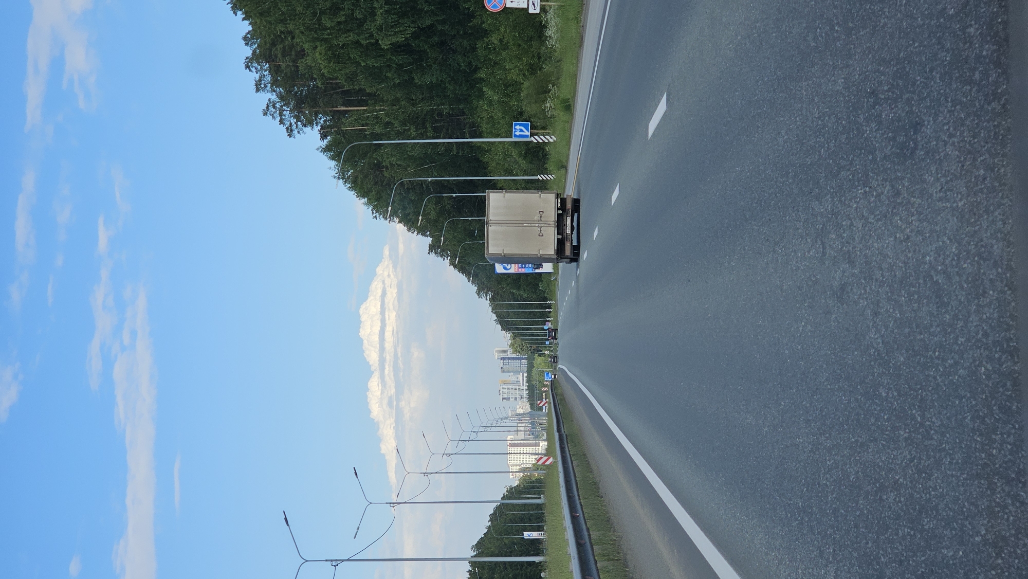 A truck travels down a highway in Yekaterinburg, Russia. The sky is blue with white clouds. Trees line the right side of the road, and streetlights are visible on both sides. Buildings can be seen in the distance.