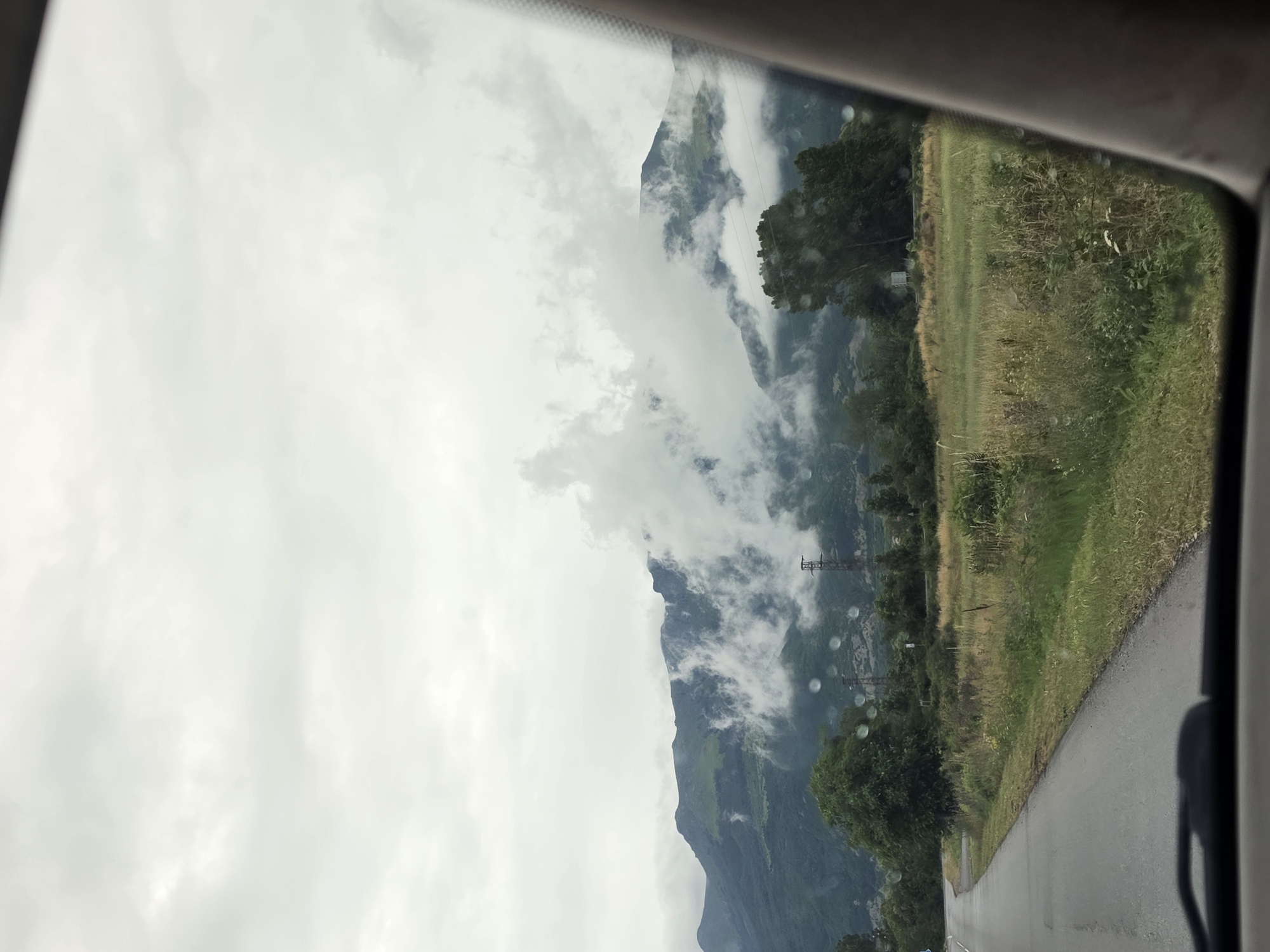 The image captures a view from inside a vehicle, looking out through the windshield. A paved road curves to the left in the foreground. To the right of the road is a grassy field with wildflowers and tall grasses. Beyond the field, several trees are visible, including a large, leafy tree to the right. In the background, a mountain range dominates the scene, with clouds obscuring parts of its peaks and slopes. Electrical towers and wires can be seen scattered across the landscape. The sky is overcast with a light gray hue. There are numerous water droplets visible on the surface of the windshield, suggesting it may have recently rained or is currently raining. No people are visible in the image. The scene appears to be a rural or semi-rural mountainous area.