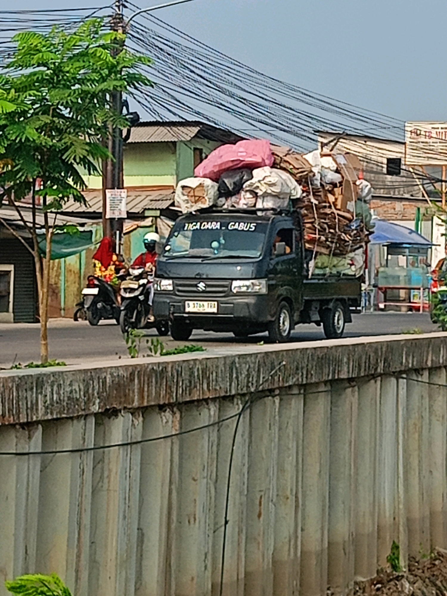 In Sriamur, Indonesia, a Suzuki Carry truck overloaded with recyclable materials, including cardboard boxes and plastic bags, is captured in motion. The truck has "TIGA DARA GABUS" written on its front. A license plate, "B 3769 TRA," is visible. Two individuals on motorcycles are also present on the road. One is wearing a red shirt and a yellow floral pattern hijab, while the other wears a red shirt and a white helmet. The scene unfolds on a street bordered by a concrete wall and a body of water, with dense overhead electrical wires and various buildings in the background. The presence of a small tree and foliage suggests an urban environment.