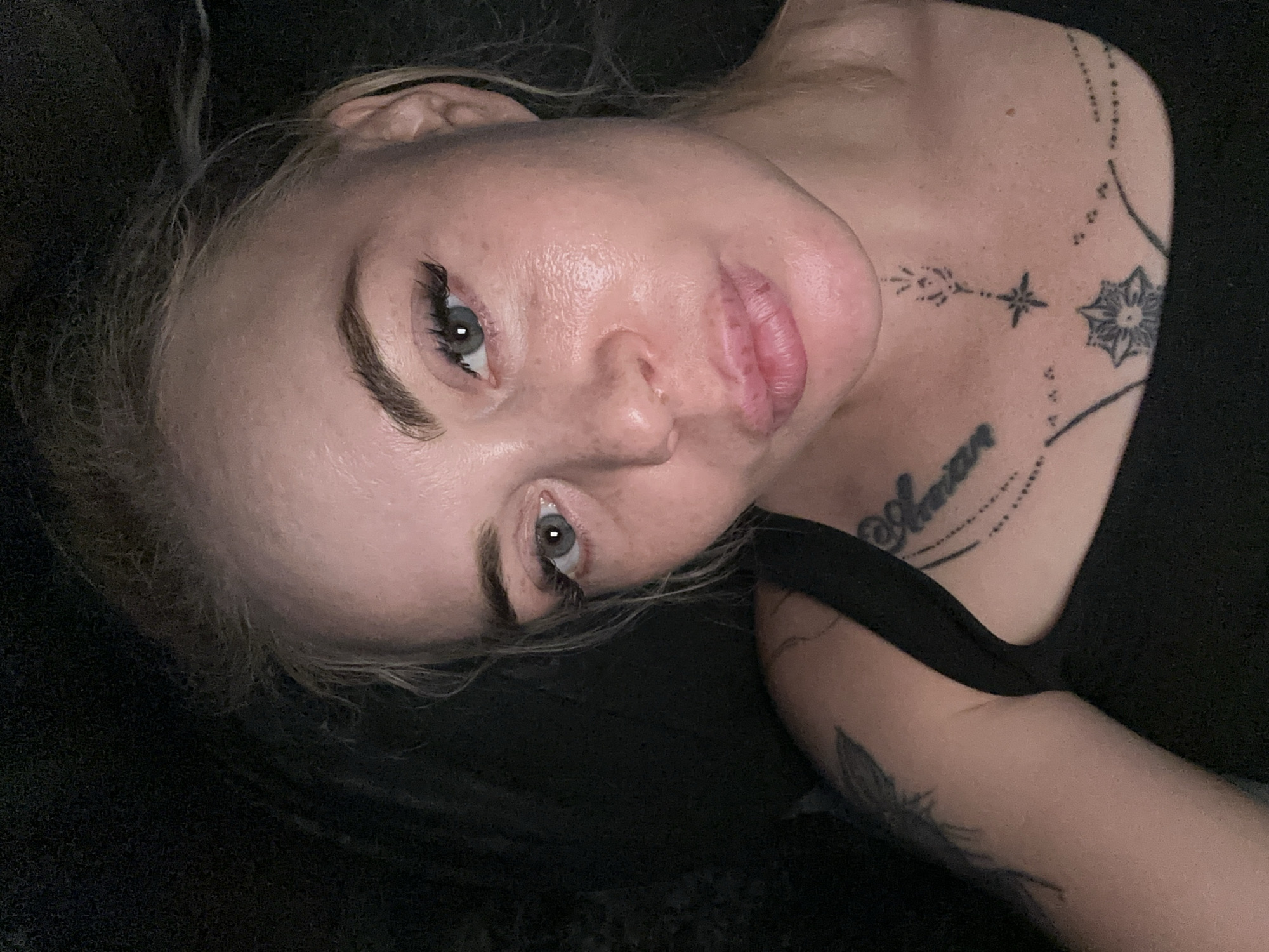 A close-up, head-and-shoulders selfie of a fair-skinned woman with blonde hair pulled back is taken in a dark, unidentifiable environment. She is wearing a black tank top, and several tattoos are visible on her chest and left shoulder. The woman's face is illuminated, revealing light makeup, including mascara and possibly eyeliner. Her eyes are open and looking towards the camera, and her lips are slightly parted. The background is mostly dark and out of focus, offering no discernible details about the location.