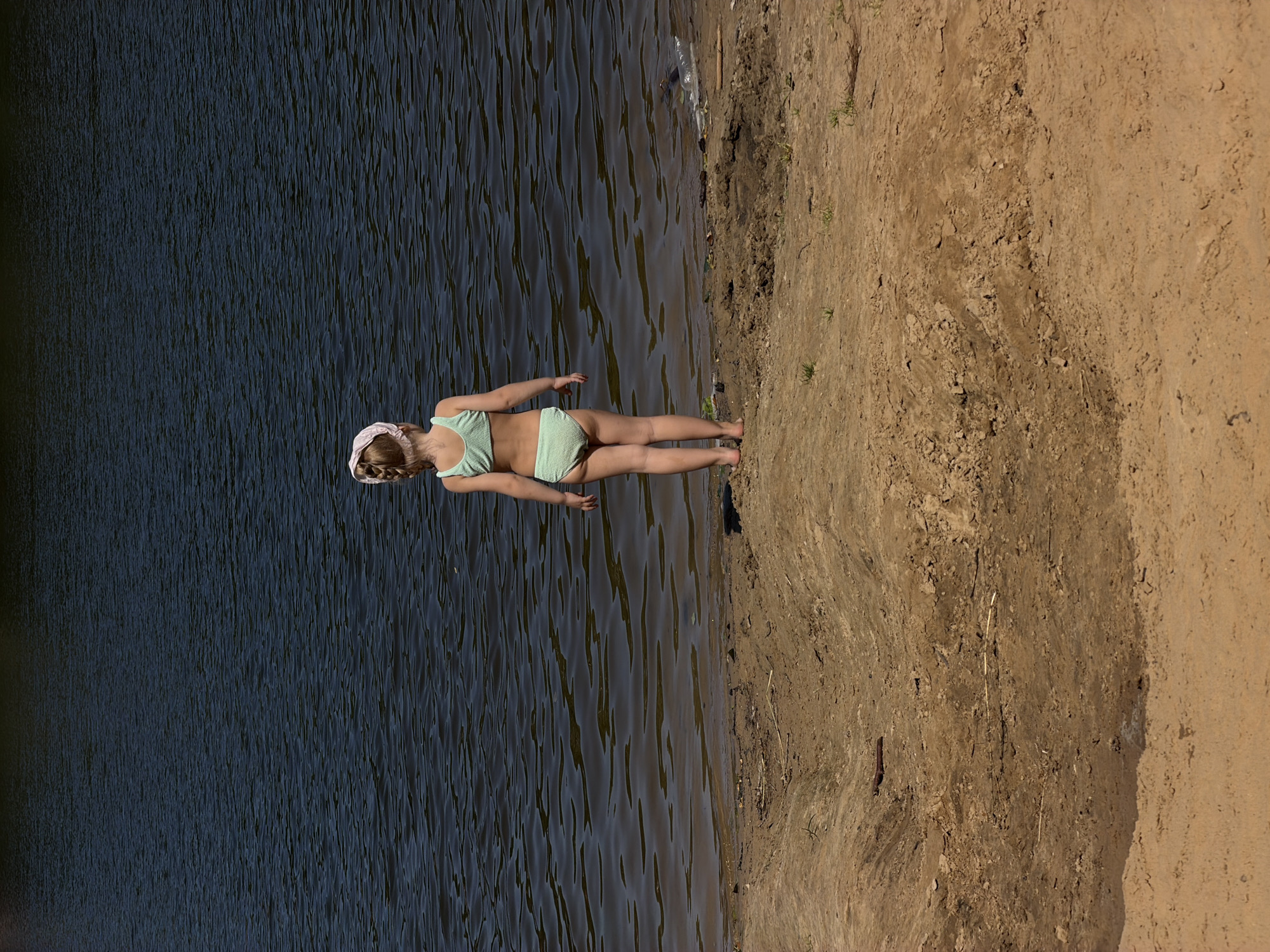In this image, a young girl is lying on the sandy shore of a body of water. She is wearing a light green bikini and a light pink headband that keeps her braided hair in place. Her body is positioned horizontally, facing towards the water, with her legs extended and her right arm bent at the elbow, her hand resting on the sand. The water, which appears to be a dark, calm lake or river, fills the left side of the frame, with ripples indicating gentle movement. The sandy shore, on the right, is uneven and textured, with sparse dry grass growing in patches. The lighting suggests it is a sunny day, casting distinct shadows and highlighting the dry earth. The scene evokes a sense of peacefulness and quiet enjoyment of a natural outdoor setting. The lack of any visible text or other objects makes the girl and the natural elements the sole focus of the image.