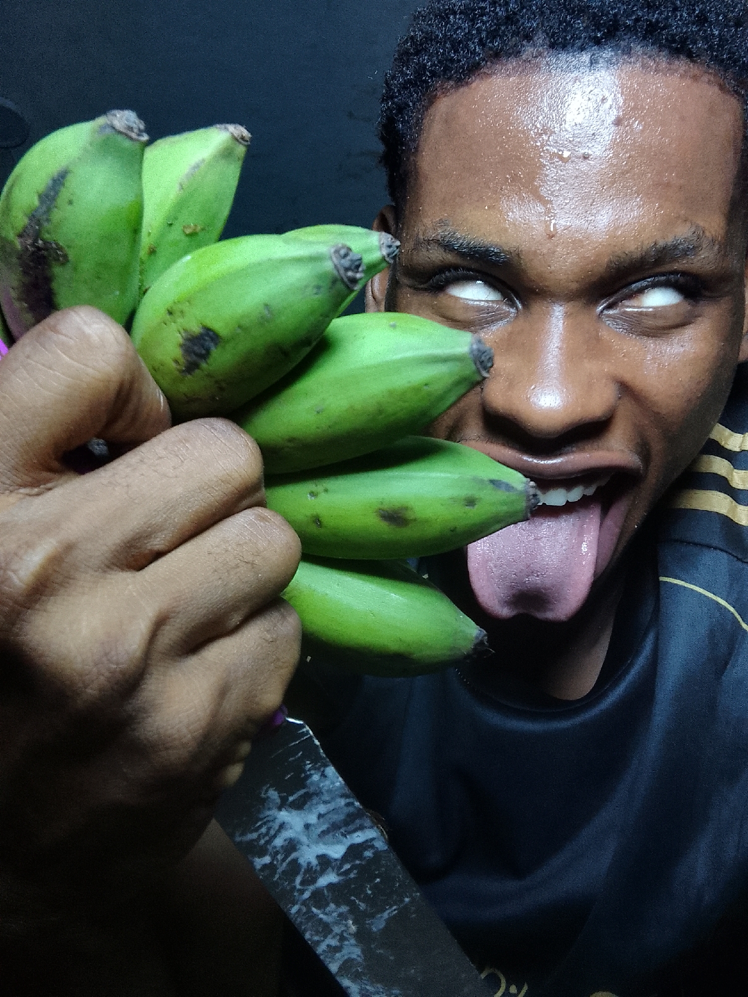 A person with dark skin, wet with sweat, is holding a bunch of green bananas in their left hand and a knife with a white-streaked blade in the lower part of the frame. The person's eyes are rolled back, showing only the whites, and their tongue is sticking out of their mouth. The person is wearing a dark shirt with gold stripes on the shoulder. The location is presumed to be Nkpor, Nigeria.