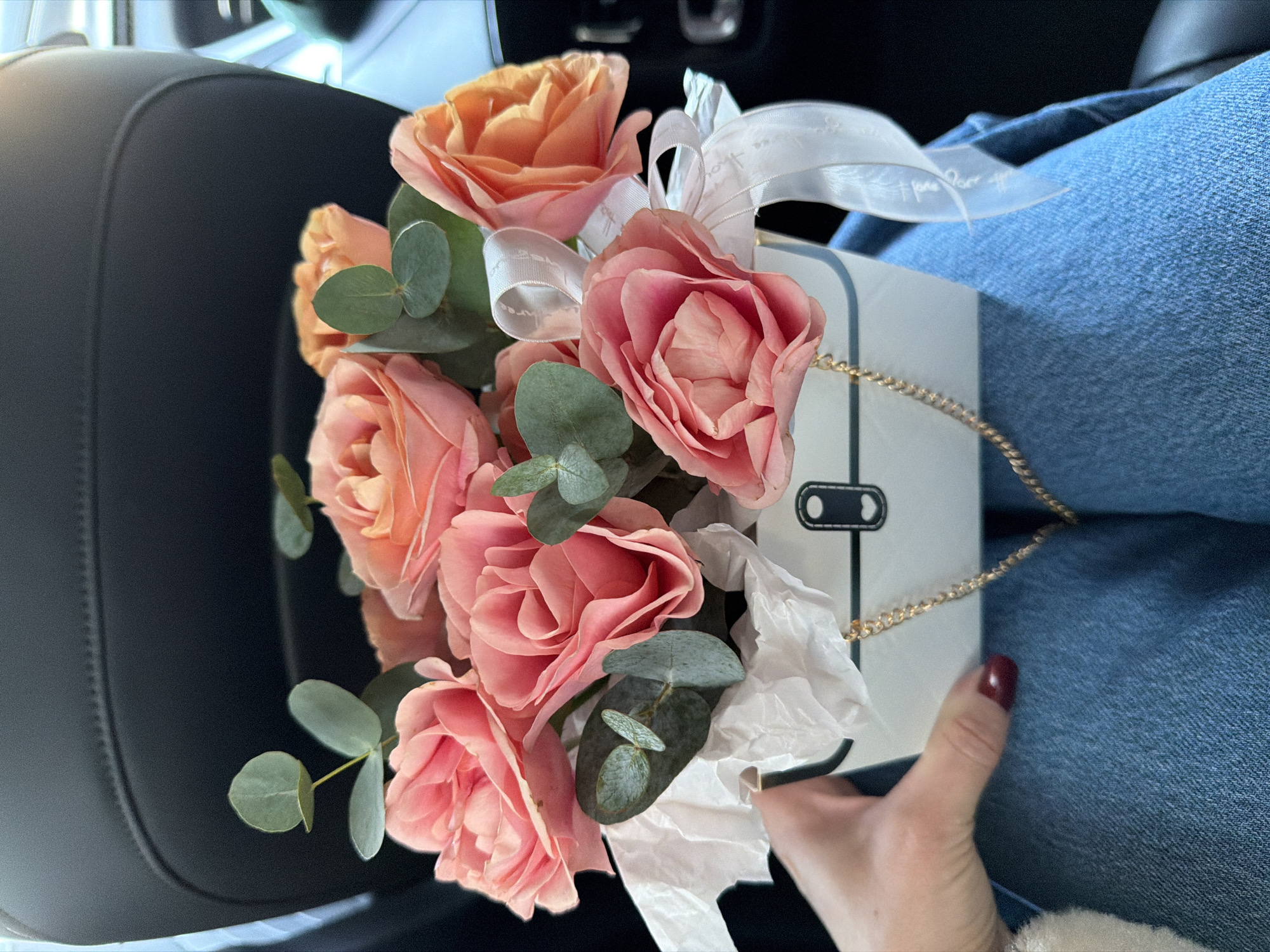 A close-up image depicts a person holding a small, decorative floral arrangement while seated, likely within a vehicle. The person's lower torso, clad in blue denim jeans, is visible, along with their left hand, featuring dark red nail polish, grasping the white, rectangular flower box.
The floral arrangement consists of approximately ten roses in shades ranging from soft pink to peach and orange, interspersed with green eucalyptus leaves. White tissue paper is visible among the flower stems within the container. The box itself is white with a black trim and features a black rectangular design resembling a lock with a white heart icon. A gold-colored chain handle is attached to the box, and a white satin ribbon, displaying the faint text "Happy New Year," is tied to its side.
The background shows the dark, upholstered seatback of what appears to be a car or similar vehicle, with a hint of light from a window on the far left. The specific location in Votkinsk, Russia, cannot be determined from the visual information provided in the image.