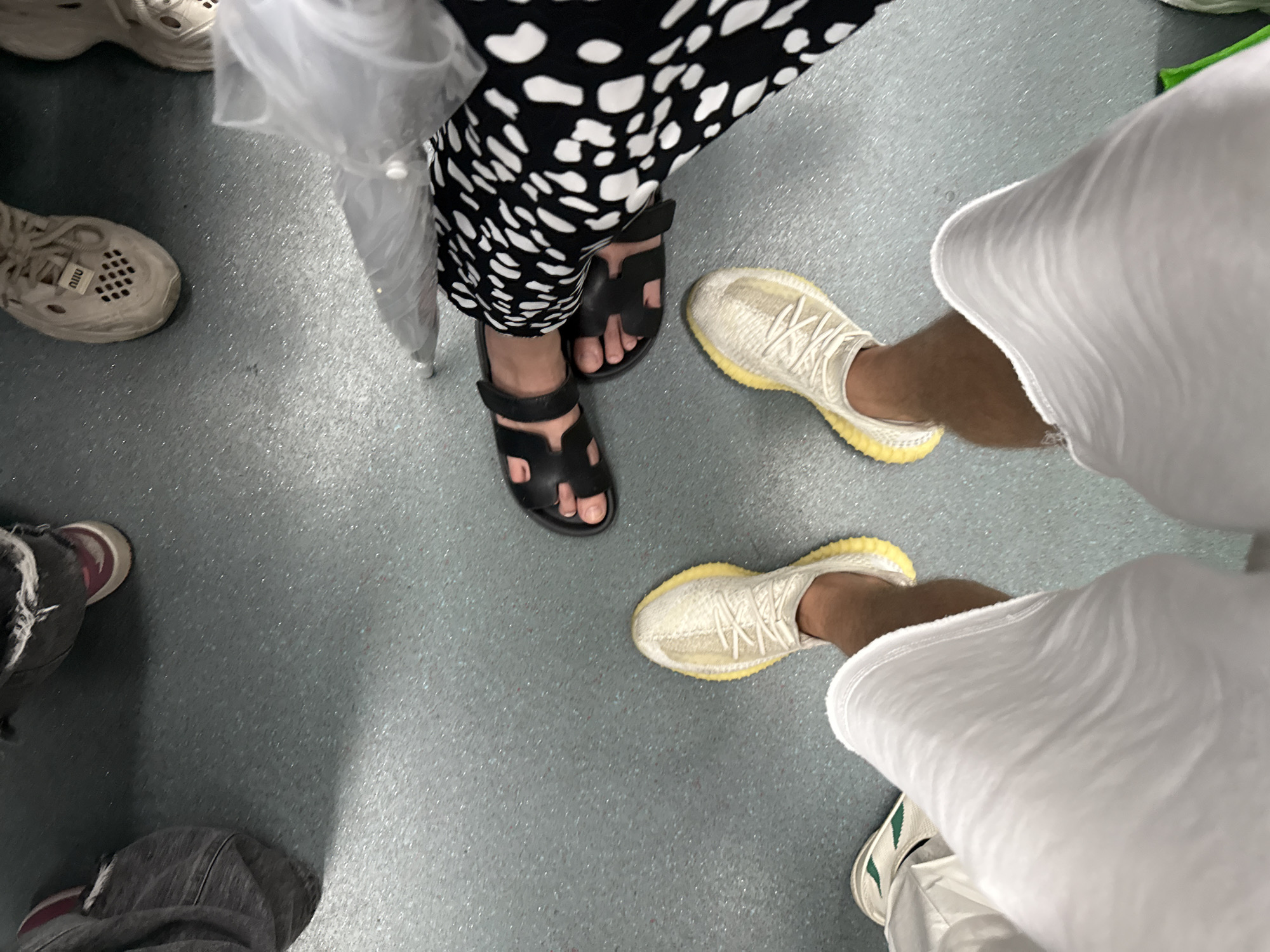 The image shows the feet of several people standing on a gray, textured floor. The floor appears to be indoors, possibly in a public space. In the foreground, a person is wearing white pants and light-colored sneakers with yellow soles. To their right, another person is wearing black sandals and black and white patterned pants. Further to the left, a person is wearing dark pants and shoes with red accents. In the background, another pair of light-colored shoes with white laces is visible. A clear plastic bag is also visible near the person in patterned pants. The image is taken from a high angle, looking down at the feet. The overall impression is of a casual gathering or transportation scenario. It is not possible to determine the specific location in Haizhu District, China, from the provided image.