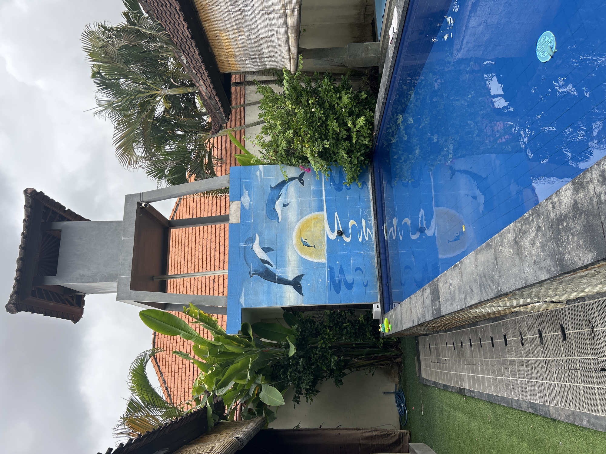 The image depicts a swimming pool area with a mural of dolphins and a sunset on a blue tiled wall. The pool itself is a bright blue, reflecting the overcast sky. To the left of the pool is a paved area with a drainage grate, and beyond that, artificial turf. In the background, there are traditional Balinese-style buildings with orange tiled roofs, lush green tropical plants, and a grey concrete tower structure. The overall scene suggests a resort or private villa in Kuta, Indonesia, given the architectural style and tropical vegetation. There are no people visible in the image.