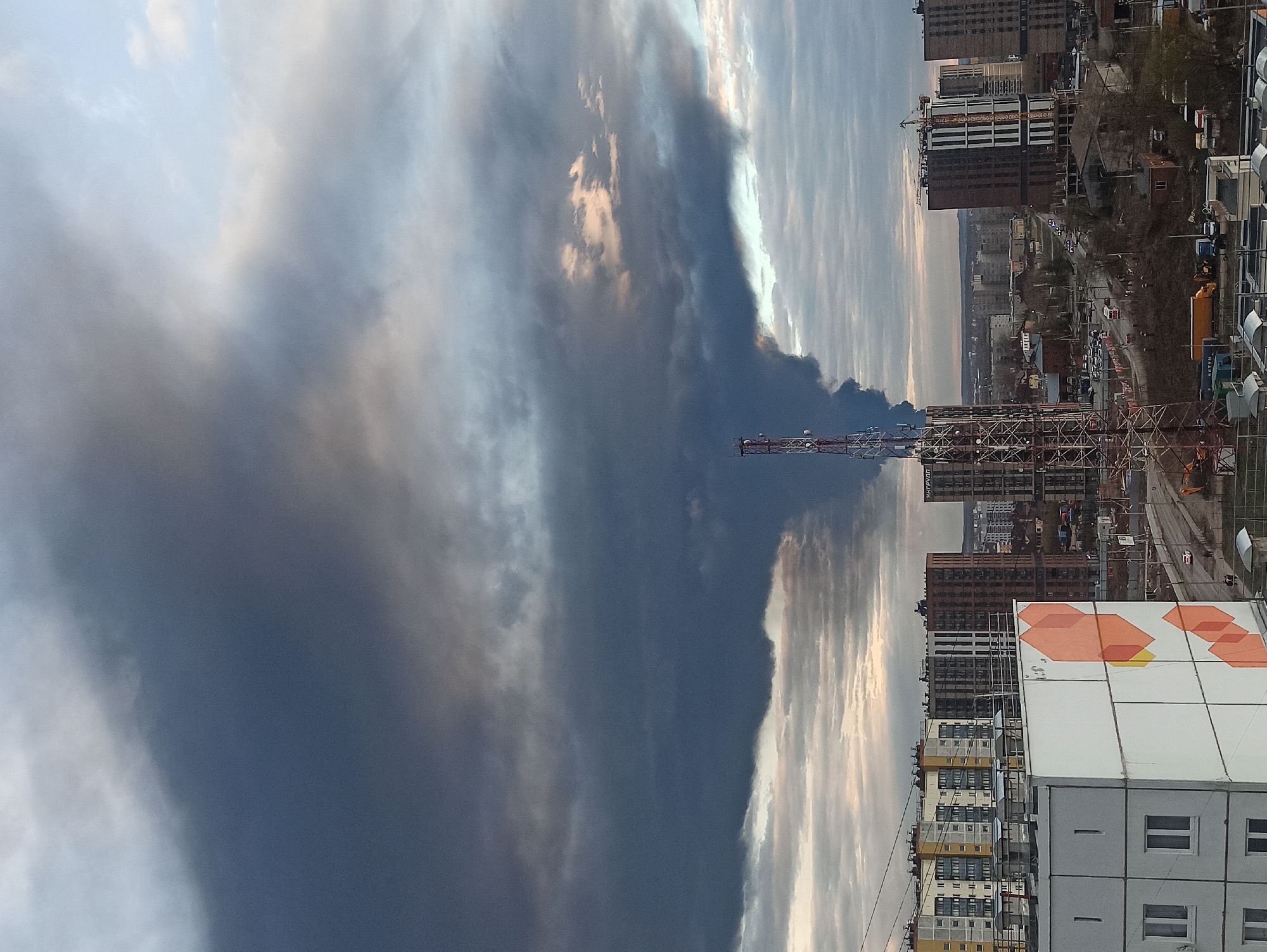 The image captures an urban landscape under a dramatic and somewhat ominous sky. A vast, dark plume of smoke or industrial emission dominates the upper left portion of the sky, contrasting sharply with patches of lighter clouds and pale blue sky illuminated by what appears to be late afternoon or early morning light towards the upper right. The density and darkness of the plume suggest a significant event such as a large fire or industrial discharge.
The cityscape below features a mix of residential and commercial structures. Several tall, dark-colored high-rise buildings are visible in the midground and background. Active construction is suggested by a prominent crane near some of these structures. A very tall, lattice-structured tower, likely for telecommunications, stands in the midground, partially obscured by the dark plume. In the foreground on the right, there is a white modular building adorned with vibrant orange and yellow geometric hexagonal patterns, adjacent to a multi-story residential building with yellow and grey facades. A road with a few vehicles is discernible between the buildings.
The overall atmosphere is somber due to the heavy smoke. Visible text includes "MUM" or "МУМ" (likely Cyrillic) on a small yellow truck or container in the midground, and "УРАЛДАН" (Cyrillic for Uraldan) on a distant building. No individuals are clearly visible.
