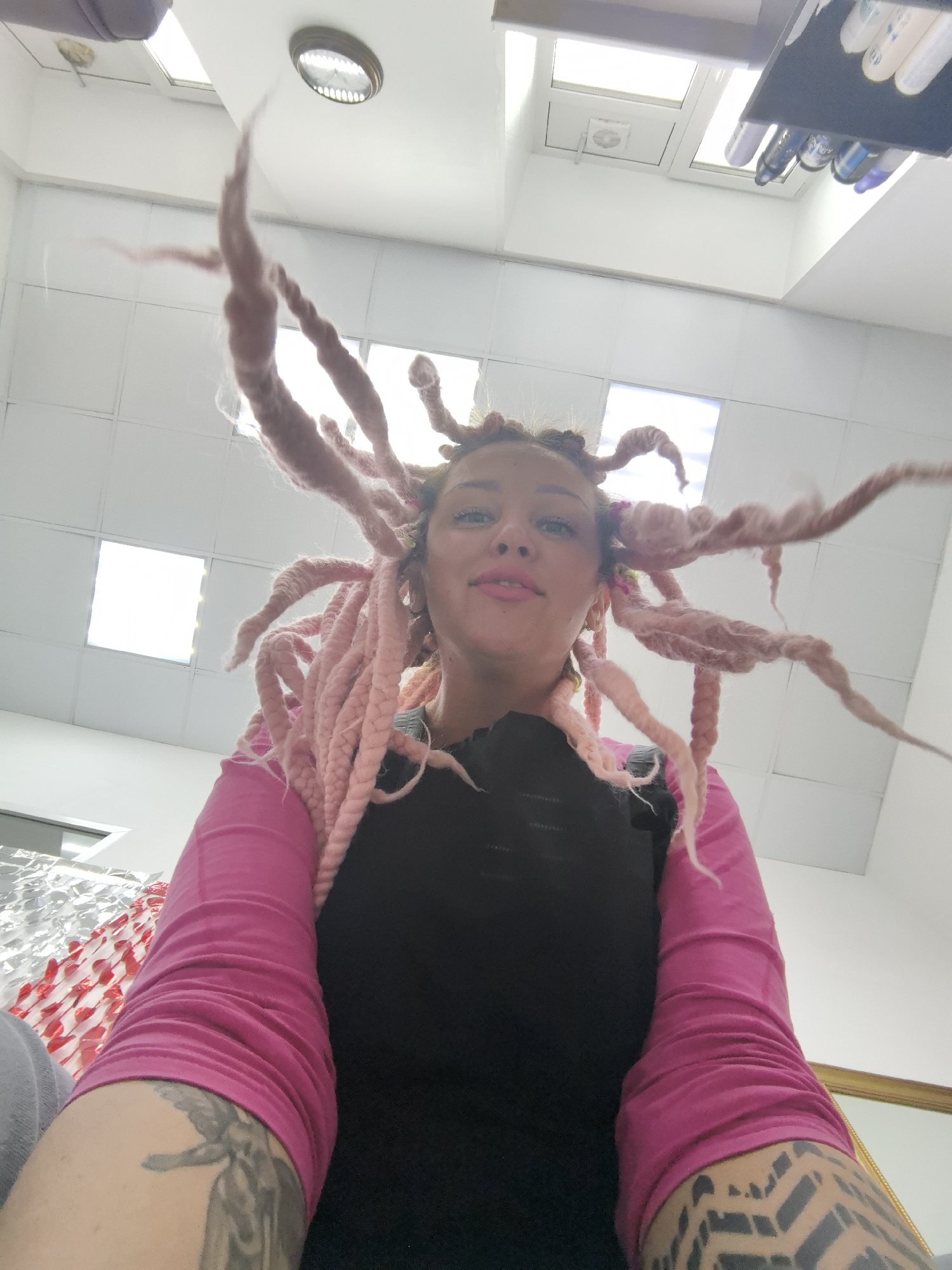 The image shows a person with pink hair in a room with white walls and a white ceiling. The ceiling has recessed lighting and a rectangular skylight. The person is wearing a black top with pink sleeves. Their arms have tattoos. The location is possibly Chaikovsky, Russia. There is not enough information to confirm.