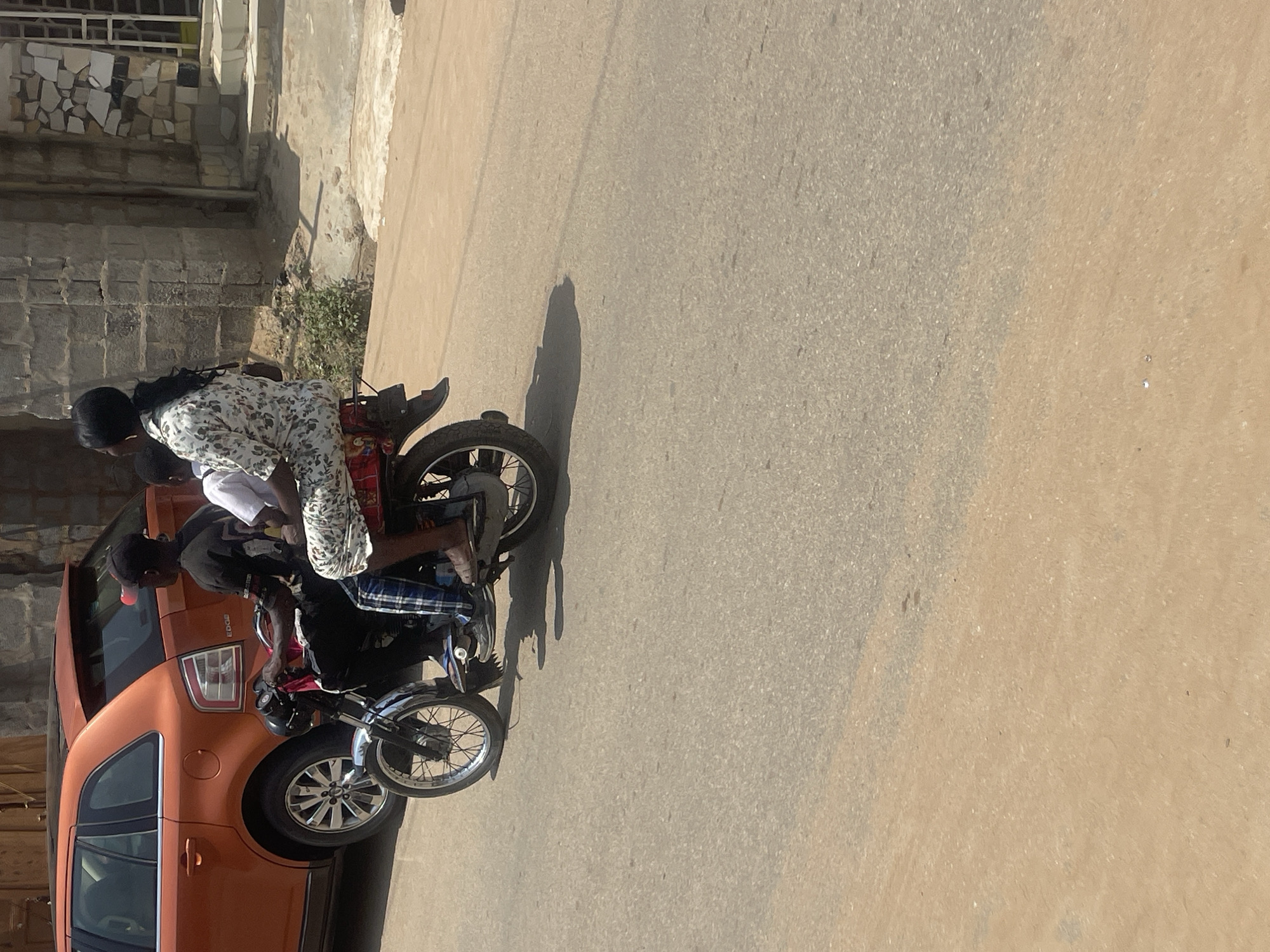 This image depicts a street scene in Abuja, Nigeria, with an orange SUV parked on the left and a motorcycle carrying three people in the center. The SUV is an older model, possibly a Ford Edge, with its rear end visible. The motorcycle is moving, with a male driver in the front, wearing a black t-shirt and plaid pants. A young boy is seated behind him, and a woman is the passenger furthest back, wearing a white patterned dress and her hair in a ponytail. The motorcycle has chrome accents on the front and rear fenders. The street is paved with asphalt and appears to be in a residential or commercial area, with buildings visible in the background. The weather is sunny, casting shadows on the ground.
