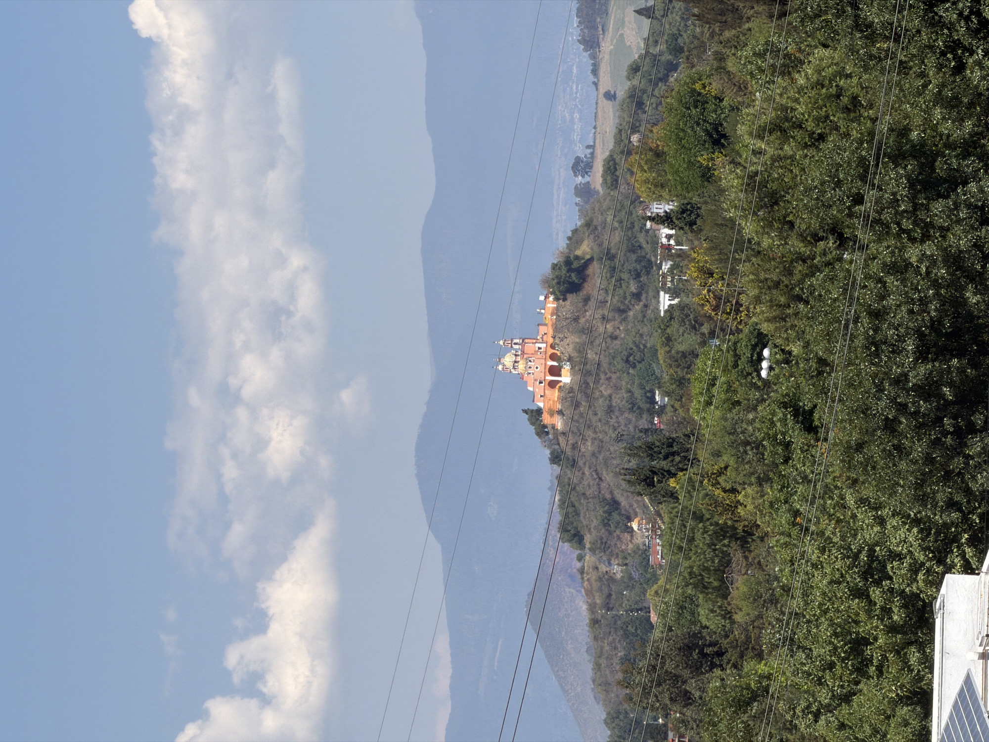 The image presents a panoramic landscape view near San Andrés Cholula, Mexico, under a bright, partly cloudy sky. A prominent colonial-style church, characterized by its vibrant light orange and yellow facade, multiple domes, and spires, stands majestically atop a green, tree-covered hill in the midground. This architectural landmark is likely the Santuario de Nuestra Señora de los Remedios, a well-known site in Cholula.
Behind the church, a series of hazy, muted blue mountains or hills recede into the distance, adding depth to the expansive scene. The foreground and midground are densely populated with lush, dark green trees and foliage, covering the undulating terrain. Scattered among the greenery, various white and light-colored buildings or houses can be seen on different hillsides. Several dark power lines traverse horizontally across the entire width of the frame, cutting across the sky, hills, and trees. In the bottom right corner, a portion of a white building or rooftop is visible, equipped with what appear to be solar panels.
The scene depicts a clear, bright daytime with scattered white clouds in a light blue sky. There is no visible human activity, creating a serene and timeless atmosphere that highlights both the natural beauty and the historical architecture of the region. No visible text is present in the image.