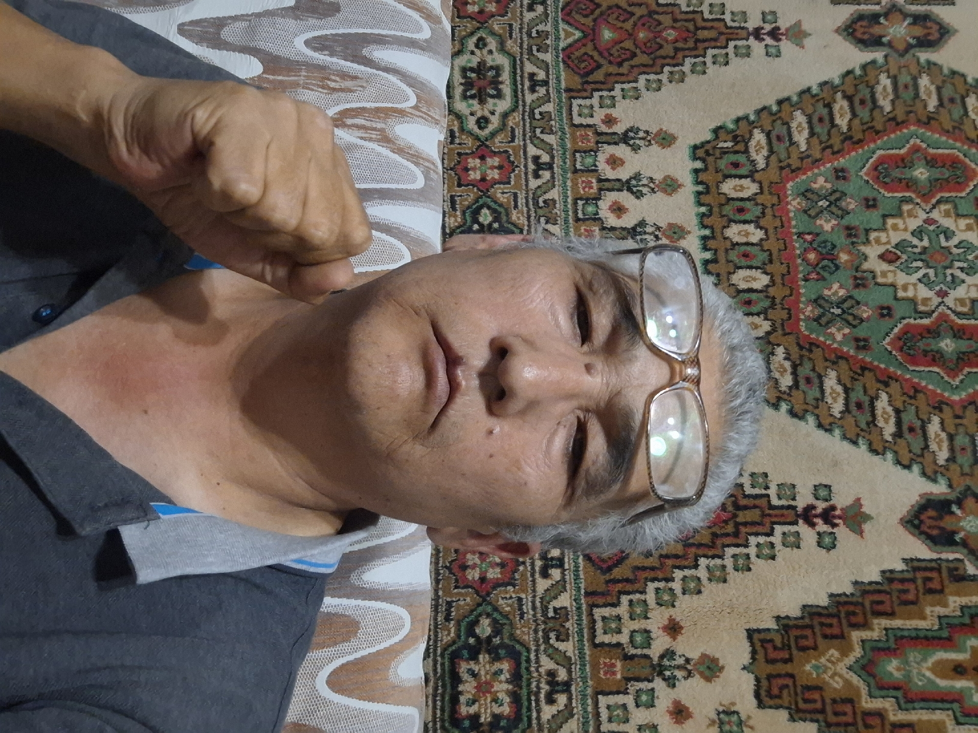 A middle-aged man with graying hair and glasses is shown in a selfie. He is wearing a dark gray polo shirt with a light gray and blue collar. His right fist is clenched and held up near his face. The background features a tapestry with intricate geometric and floral patterns in shades of red, green, brown, and beige, which is a common decorative element in homes in Almaty, Kazakhstan. The man is sitting on a couch with a patterned fabric. The lighting suggests an indoor setting, possibly a living room.