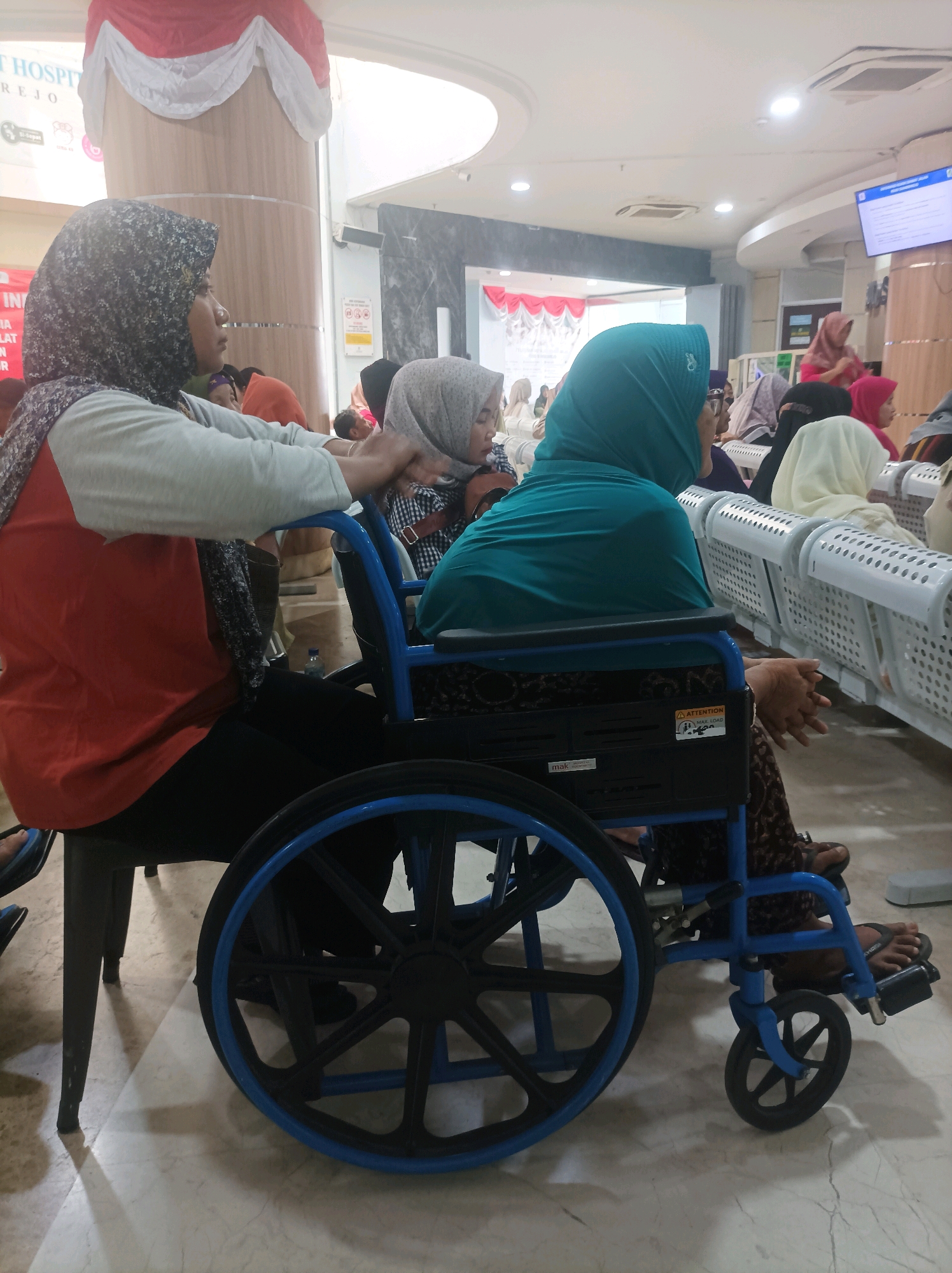 A group of people, some wearing hijabs, are waiting in what appears to be a hospital or clinic waiting area in Sumuragung, Indonesia. In the foreground, a person in an orange shirt and black pants is pushing a blue wheelchair occupied by another person wearing a teal hijab. The wheelchair has black wheels with blue rims. In the background, several rows of white plastic chairs are visible, with some individuals seated and others standing. A large screen displaying information is mounted on the wall. The floor is tiled with a light marble pattern. The environment appears clean and well-lit, suggesting an indoor public space.