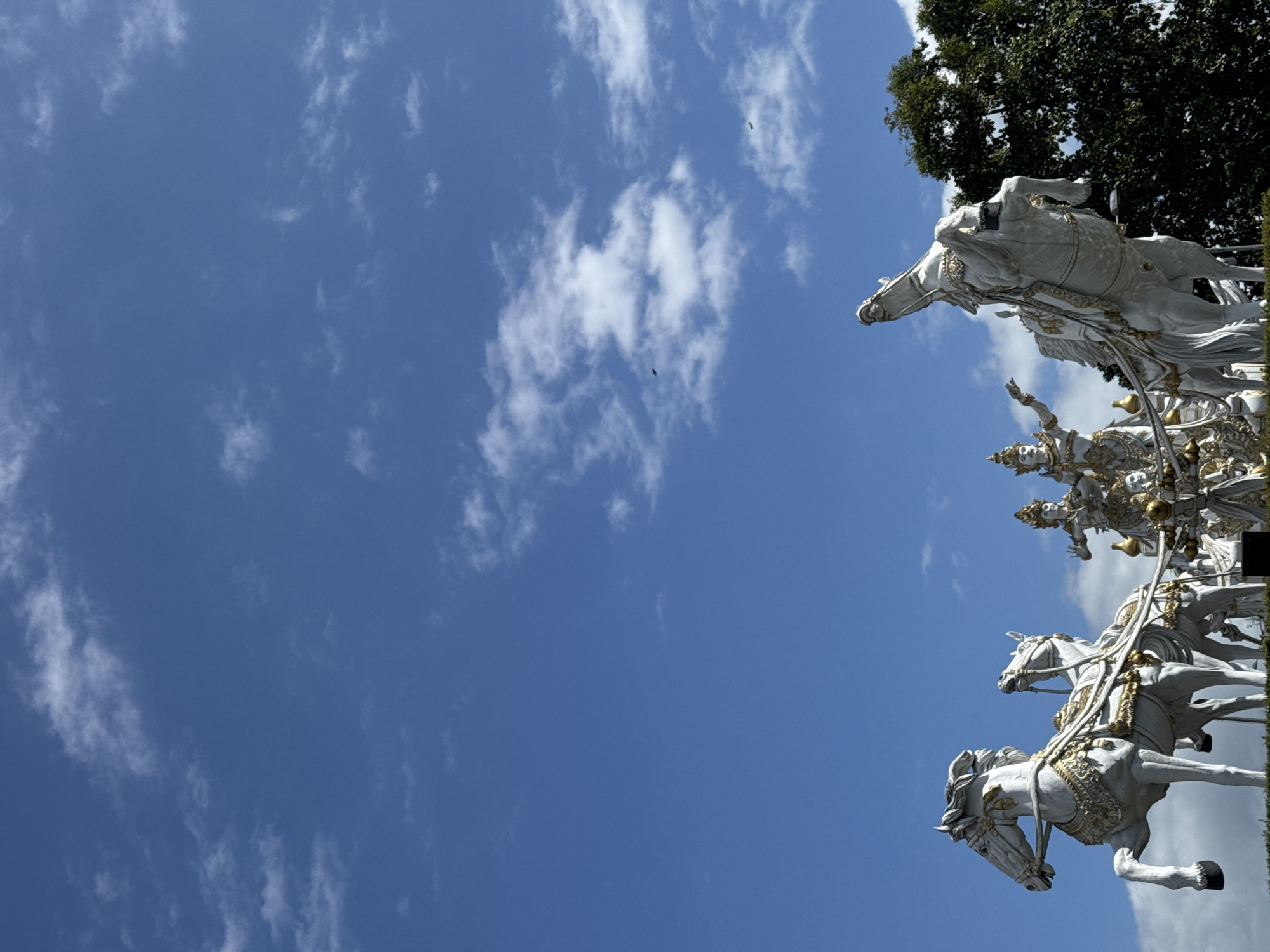 A low-angle shot reveals a statue of a chariot pulled by four horses, partially obscured by a tree. The statue, white with gold details, is set against a blue sky with scattered clouds. The statue is located in Mengwi, Indonesia.
