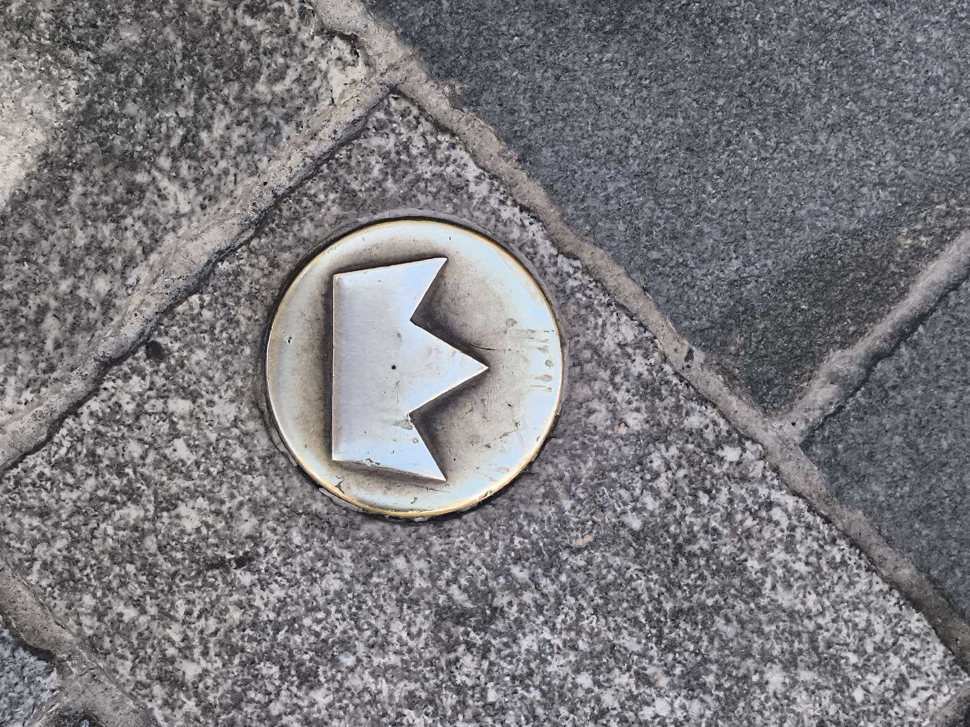 The image shows a metallic directional marker embedded in a stone pavement in Bratislava, Slovakia. The marker is circular with a stylized arrow pointing to the left, formed by three sharp angles. The marker is silver in color with a brass-toned edge. The pavement is made of grey granite tiles. The marker is likely part of a historical or tourist trail. The image is taken from a high angle, looking down at the marker. The lighting suggests an outdoor environment.
