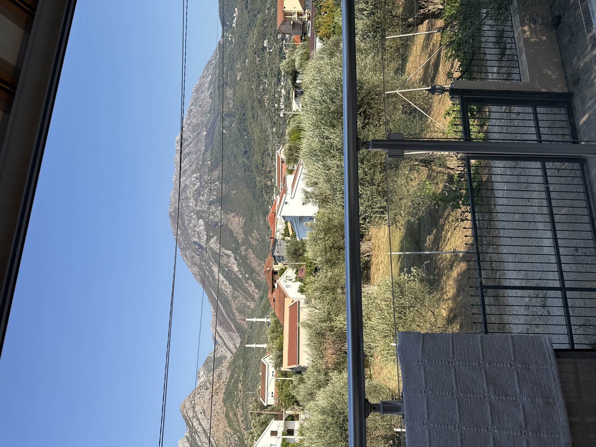 The image captures a scenic view of the town of Čeluga, Montenegro. In the foreground, a modern balcony railing with glass panels and metal posts overlooks a garden with olive trees and a black metal fence. Beyond the garden, the town unfolds with several houses featuring terracotta roofs and white walls, some with multiple stories. Two prominent minarets, characteristic of a mosque, rise above the residential buildings, indicating a significant religious presence. In the background, a large, rugged mountain range dominates the horizon, its slopes covered in green vegetation and rocky outcrops. The sky above is a clear, vibrant blue, suggesting a sunny day. Power lines are visible stretched across the frame, adding a utilitarian element to the otherwise natural and tranquil landscape. A chair with a patterned grey cover is positioned on the balcony, suggesting recent human activity or preparation for occupancy.