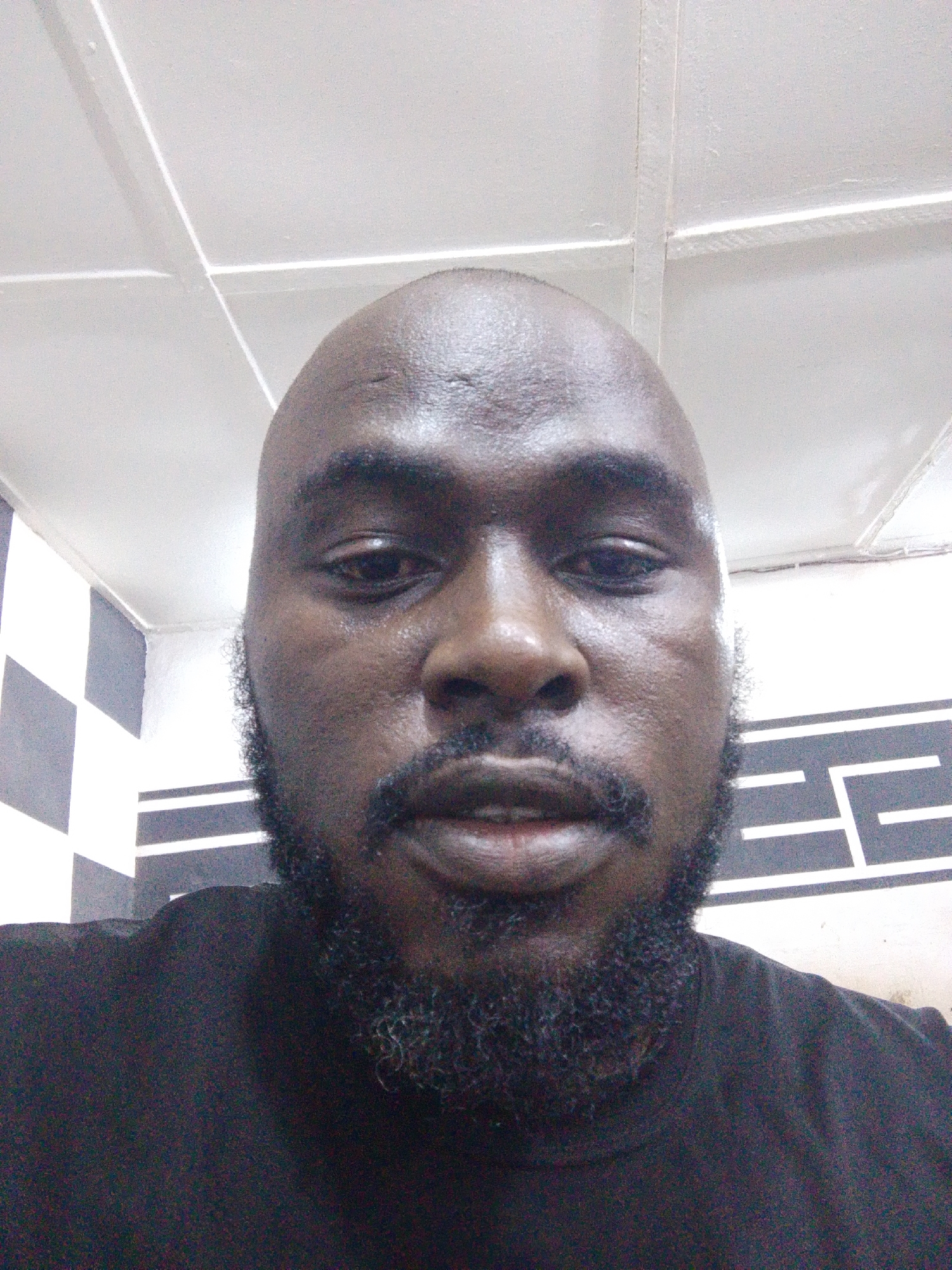 A close-up self-portrait of a dark-skinned man with a shaved head and a short, full beard and mustache. His skin appears to be moist or slightly sweaty. He is wearing a black shirt. The background shows part of a white ceiling with visible grid lines and a wall with a patterned design of alternating dark and light squares or rectangles. The exact location is indiscernible, but information was provided that it is in the city of Jos, Nigeria. No specific actions or interactions are evident.