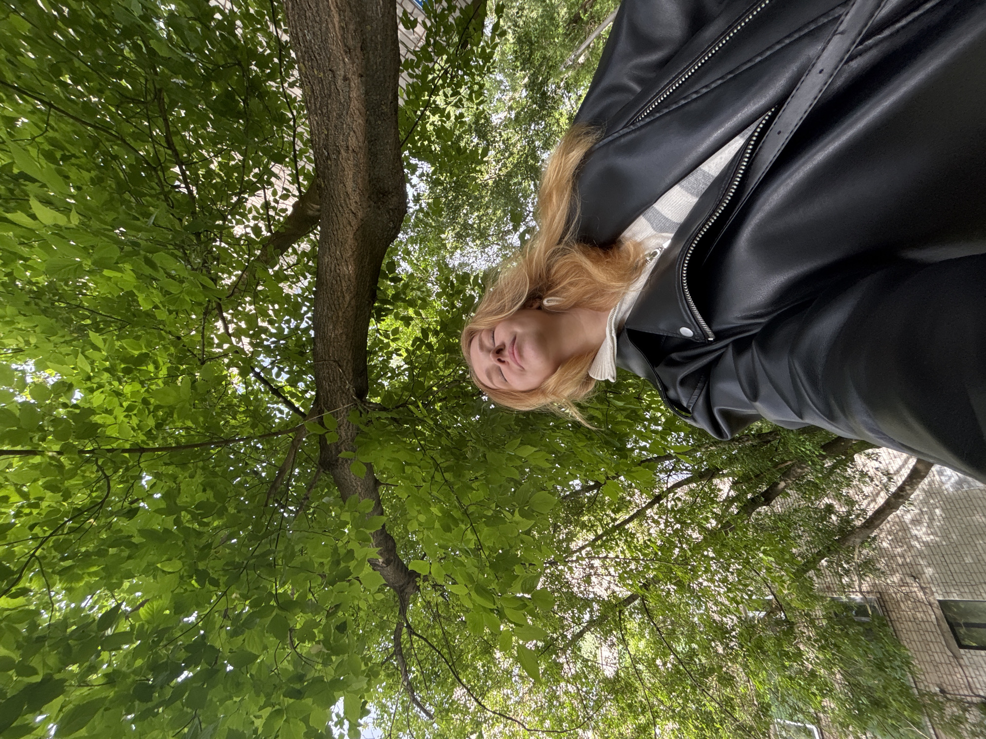 The image depicts a woman in a black leather jacket standing beneath a tree with dense green foliage. The perspective is from below, looking up at the woman and the tree canopy. The location is likely outdoors in an area with trees and buildings in the background. The presence of foliage indicates a park, wooded area, or a tree-lined street. The woman is facing downwards with her eyes closed. Her blonde hair hangs loosely, and she is wearing a light-colored top or shirt beneath the leather jacket. The specific location in Podosenikha, Russia cannot be determined.
