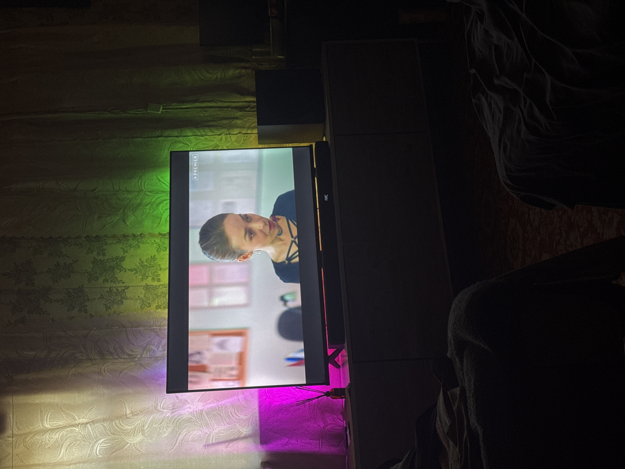A television screen displays a woman's face in an indoor setting. The screen is mounted against a patterned wallpaper illuminated by ambient light that transitions from green at the top to purple at the bottom. The television is positioned on a stand next to a tall, dark-colored storage unit. A portion of a dark blanket or clothing is visible on the right side of the frame. The image lacks specific details to determine the exact location within Makiivka, Ukraine, or to provide information about any ongoing actions or interactions.